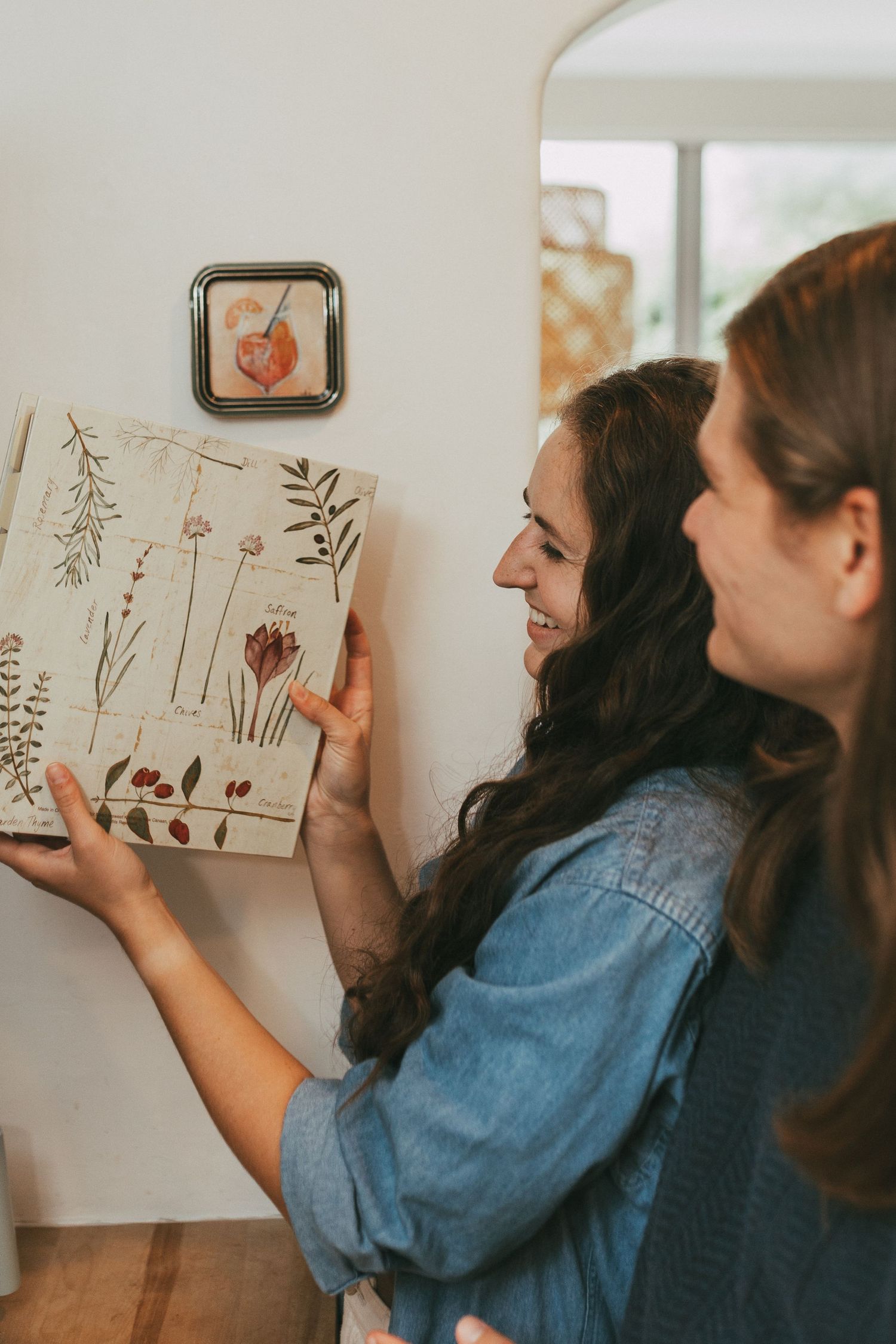 Two women smiling while hanging floral botanical art print on white interior wall.