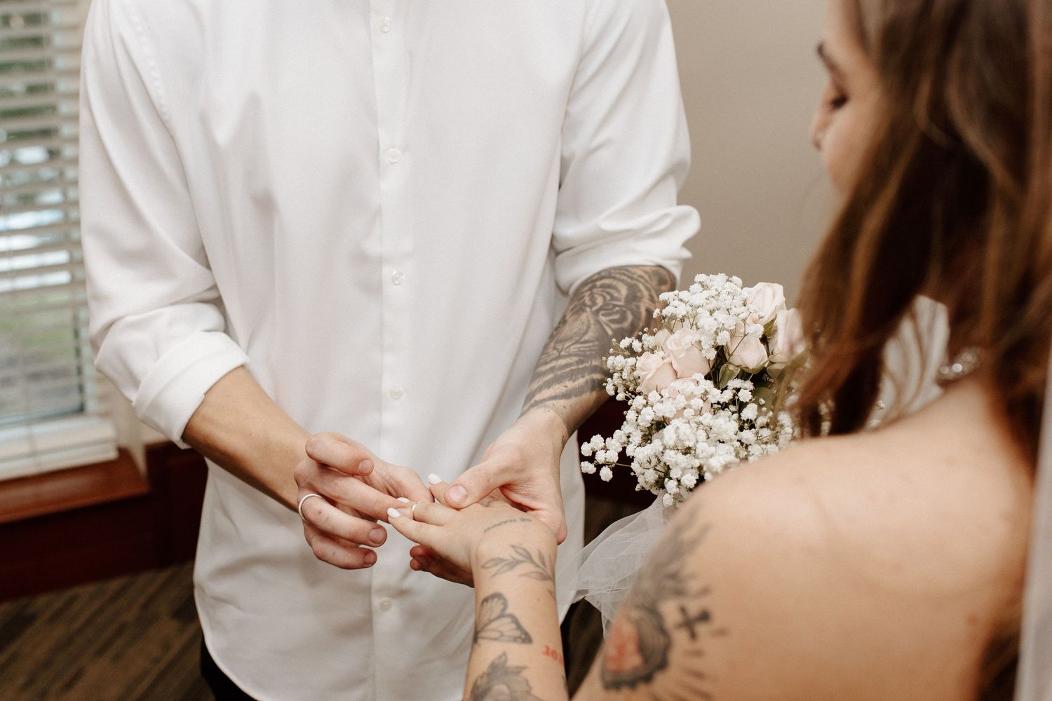 Tattooed couple exchanging rings indoors, bride holding small white and pink floral bouquet during ceremony.