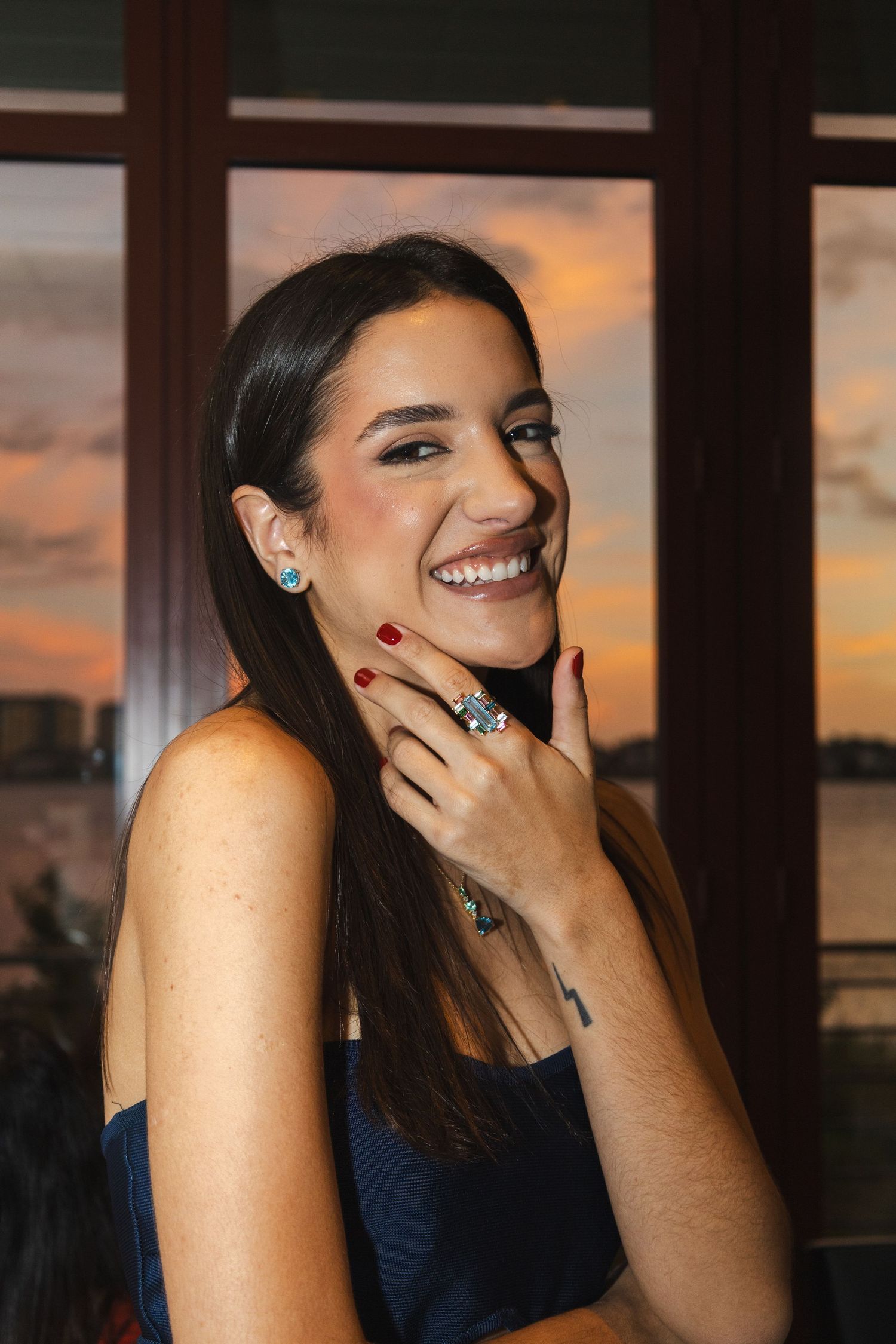 Smiling brunette woman wearing blue dress and silver jewelry with red nails at sunset.