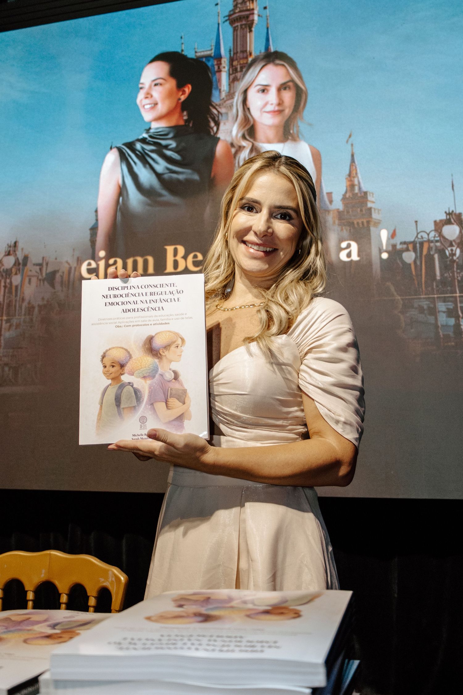 Woman in white dress holds a book at a signing event with a castle-themed backdrop featuring two portraits.
