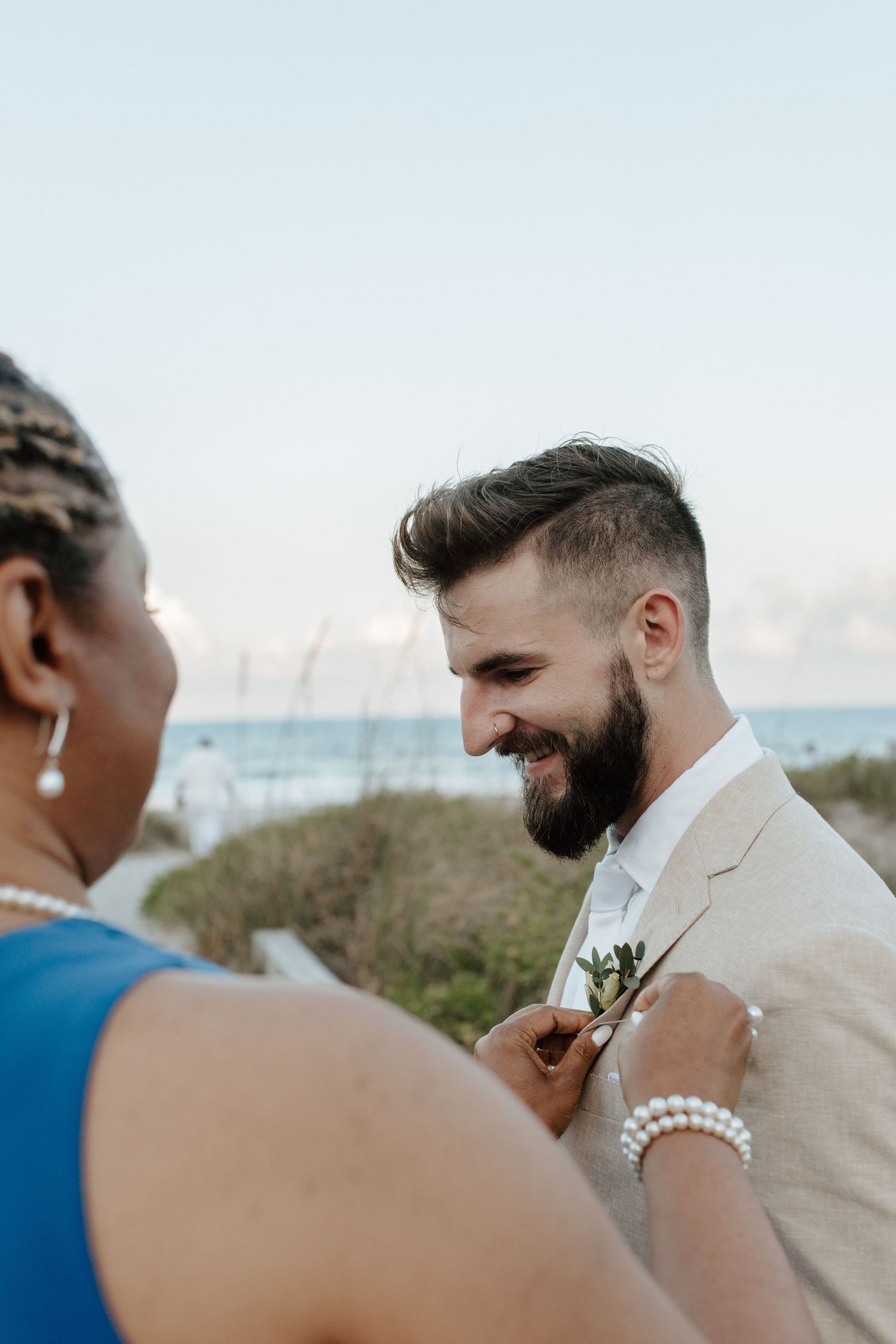 Smiling bearded groom in linen suit gets boutonniere pinned by woman in blue dress at beach wedding.