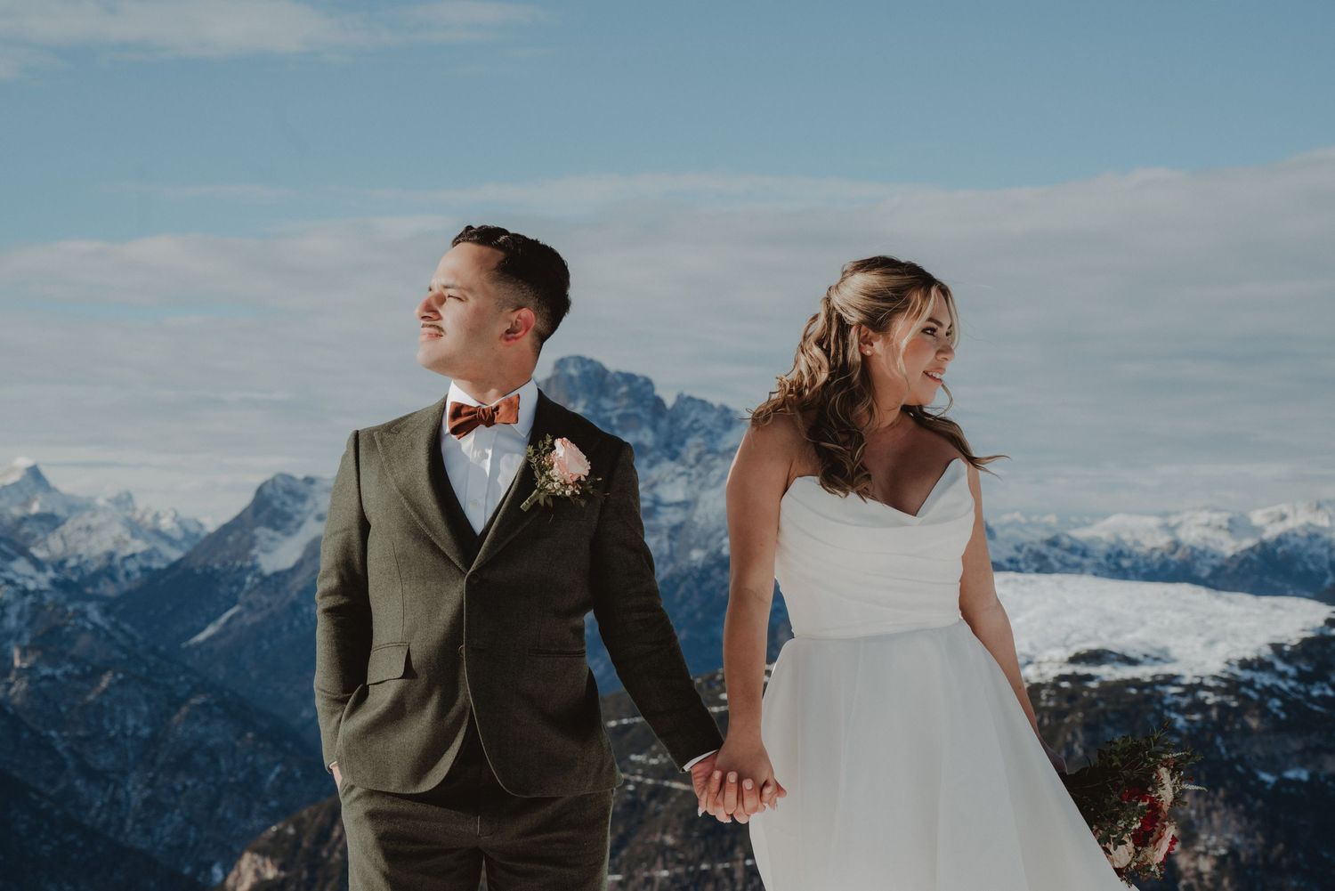 Newlywed couple holding hands and looking in opposite directions, with the snow-capped Italian mountains between them.