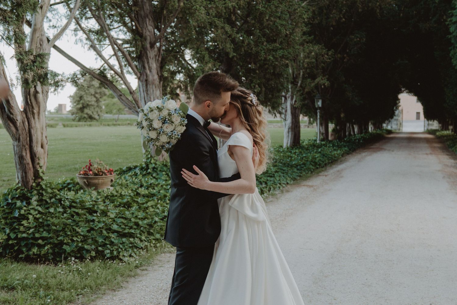 Newlywed couple kissing beneath a lush green garden archway on their wedding day.