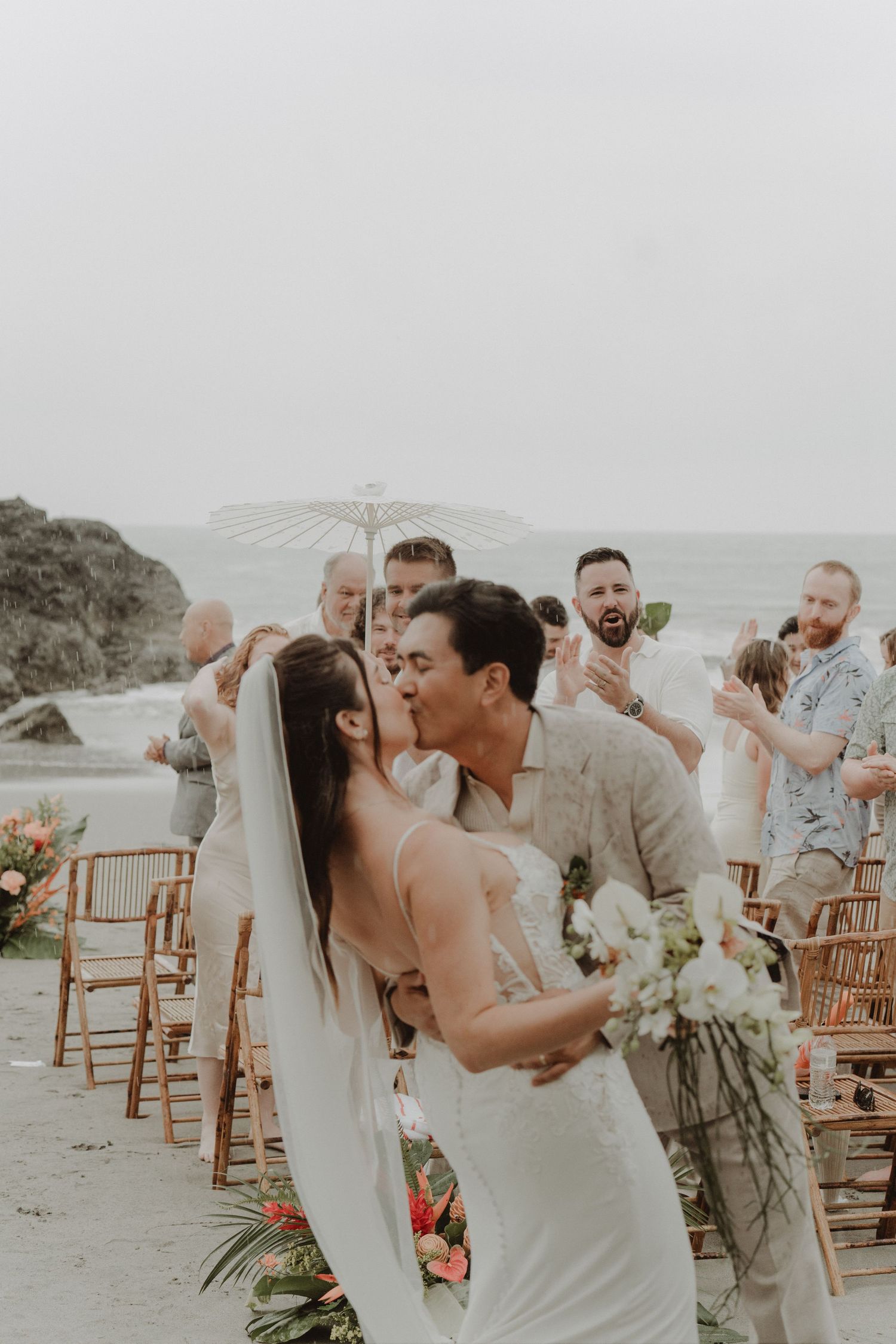 Newlywed couple kissing at the end of their beach wedding ceremony, with their guests standing behind them.