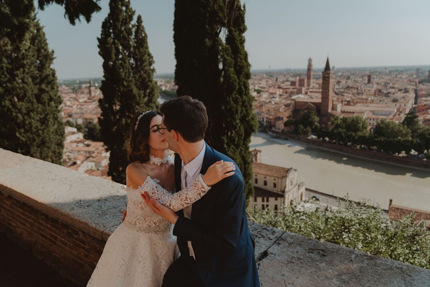 Newlywed couple kissing with a panoramic view of Verona's historic city center from above.