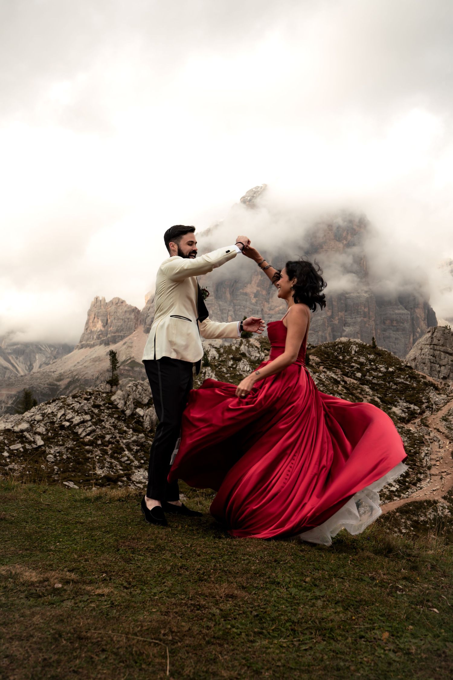Bride in a red wedding dress twirling with her groom against the backdrop of the Dolomites, Italy.