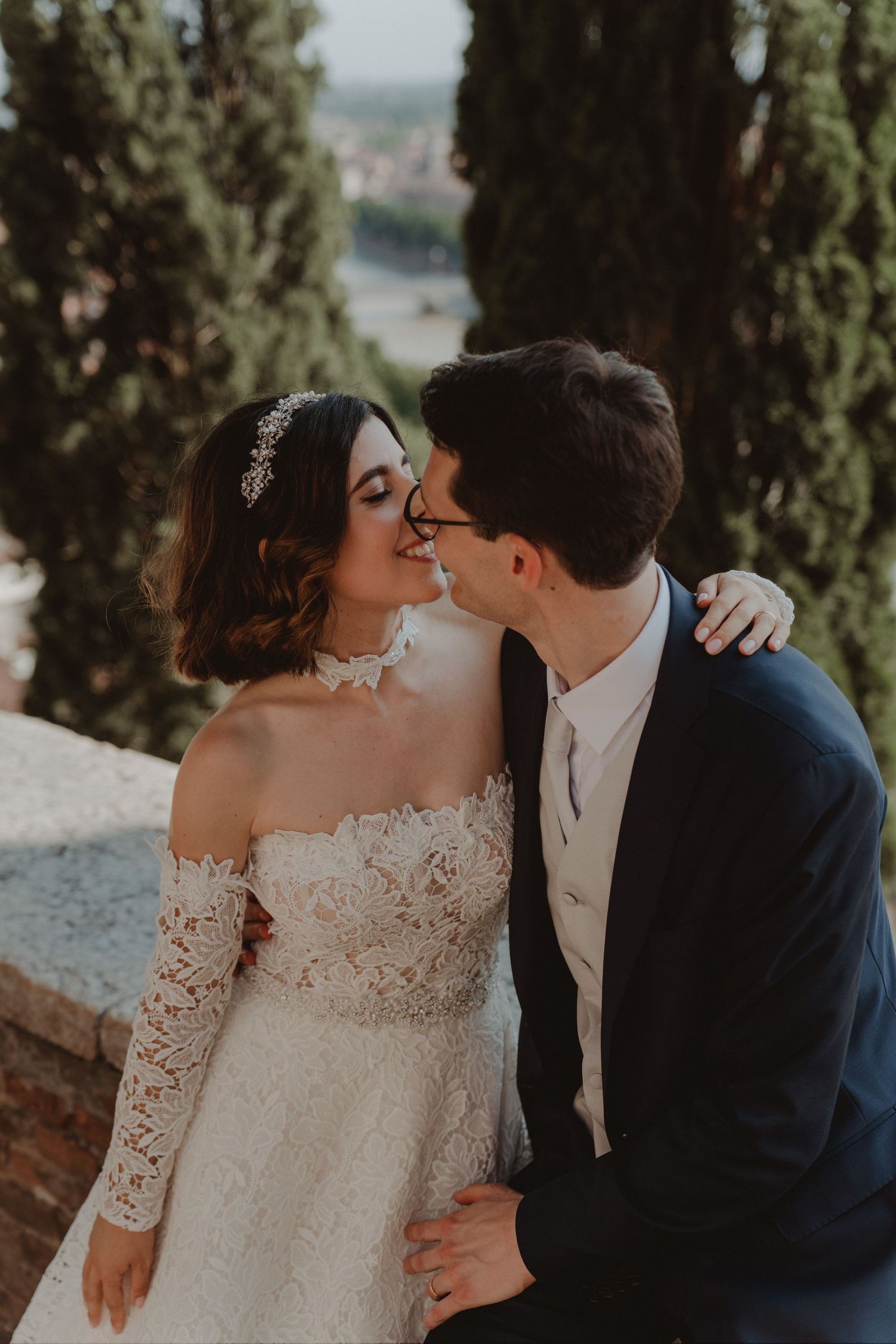 Newlywed couple kissing with a panoramic view of Verona's historic city center from above.
