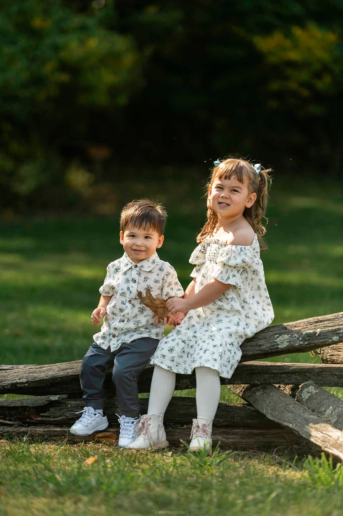 Two young siblings in matching floral outfits sit together on a rustic wooden fence in a sunny green park.