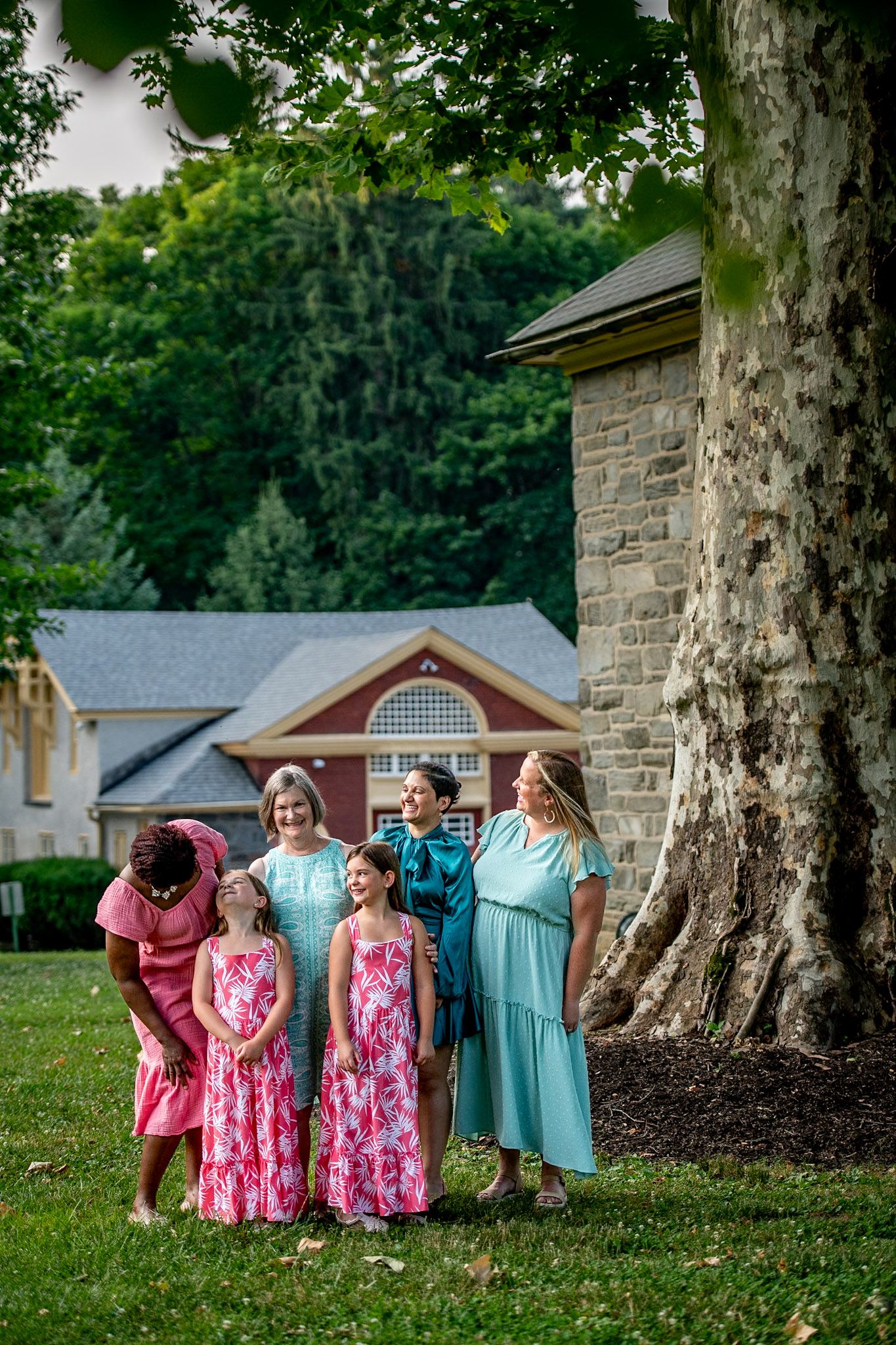 Group of women and girls in pink and teal dresses posing outdoors near a large tree and stone building.