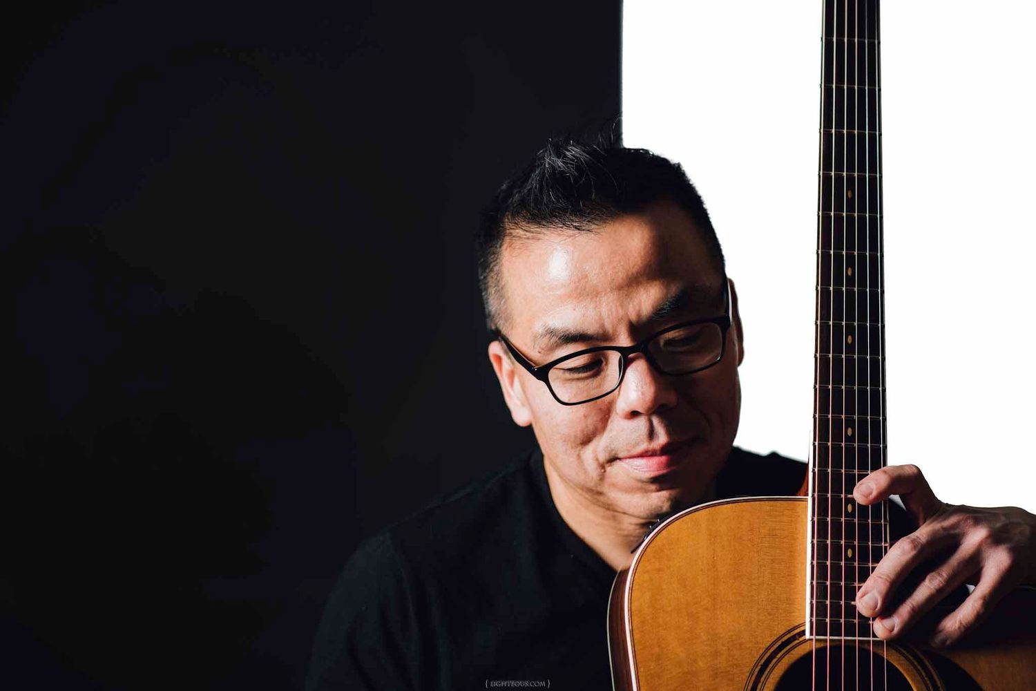 Asian man in black shirt holding acoustic guitar, wearing glasses, posed against dark background.