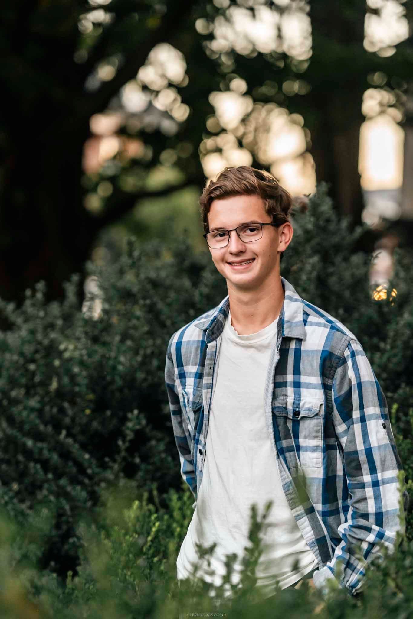 Smiling teenage boy wearing glasses and plaid shirt standing outdoors among green bushes at golden hour.
