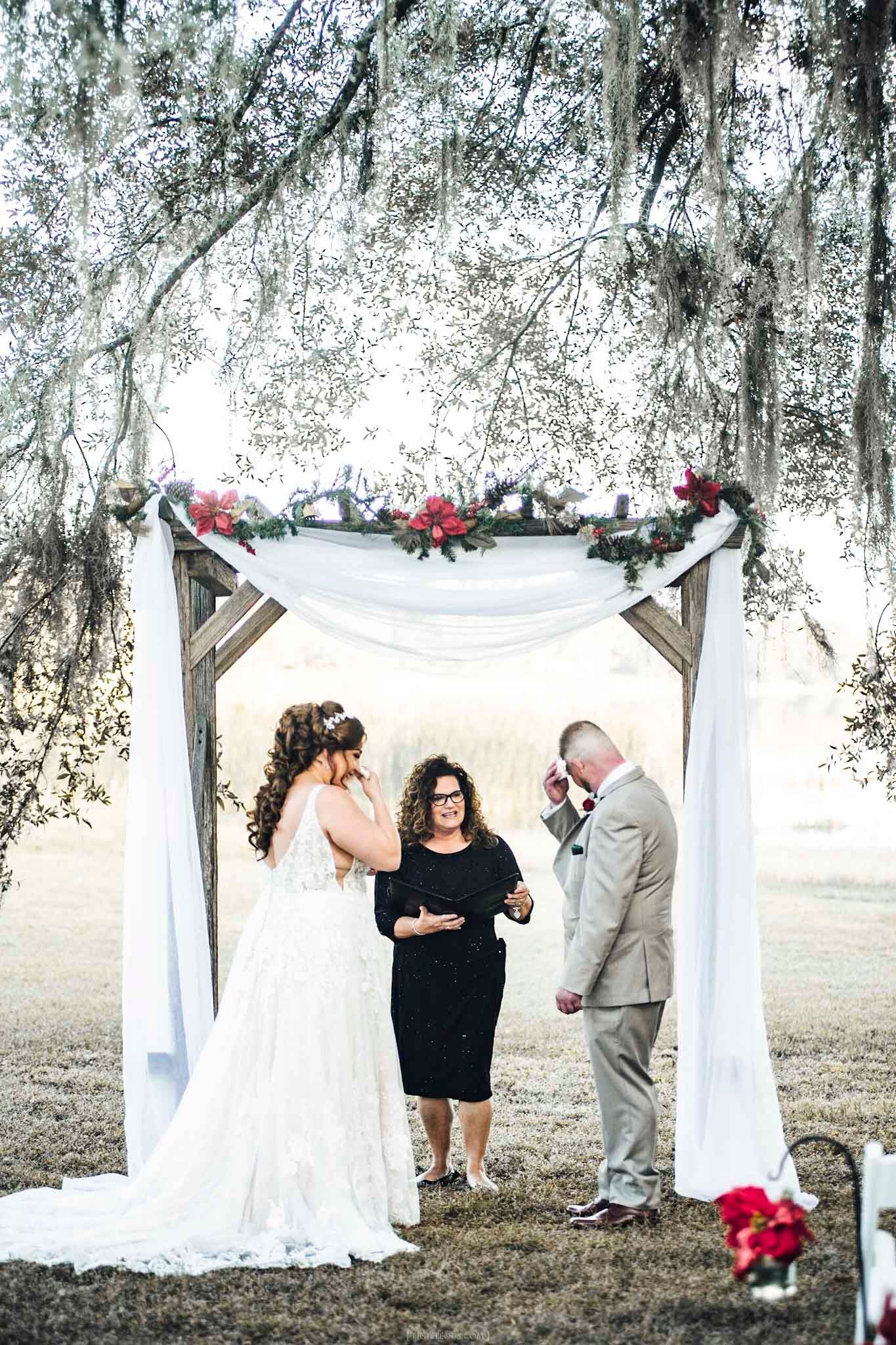 Bride and groom exchange vows under a rustic wooden arch draped in white fabric and red flowers outdoors.