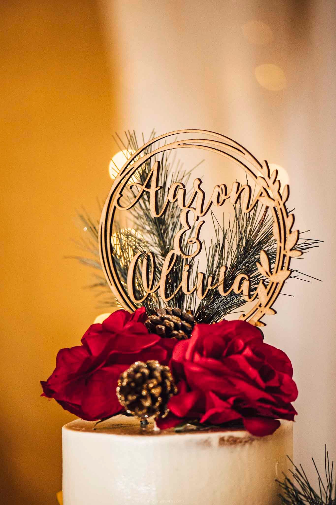 Wooden laser-cut wedding cake topper with Aaron and Olivia names, adorned with red roses and pine cones.