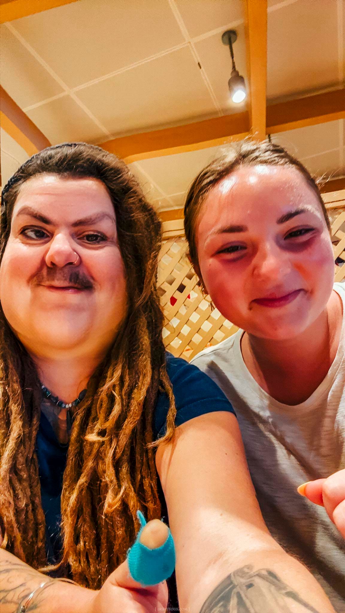 Two friends smiling together for a selfie indoors, one with dreadlocks and a mustache, the other in a white shirt.