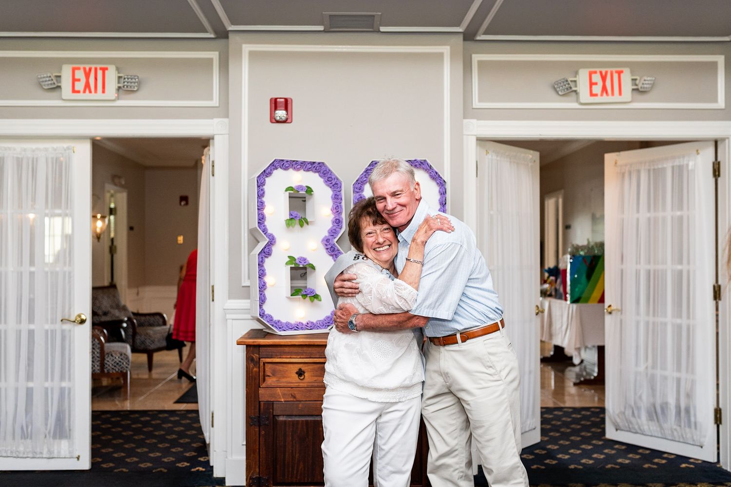 Couple hugging and smiling in front of an 80th birthday decoration at an indoor venue.