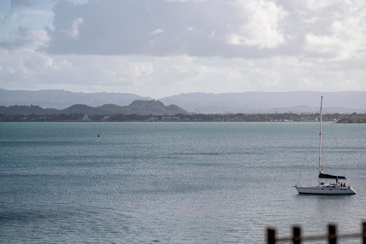 A white sailboat anchored on calm blue waters with misty green mountains in the background under cloudy skies.