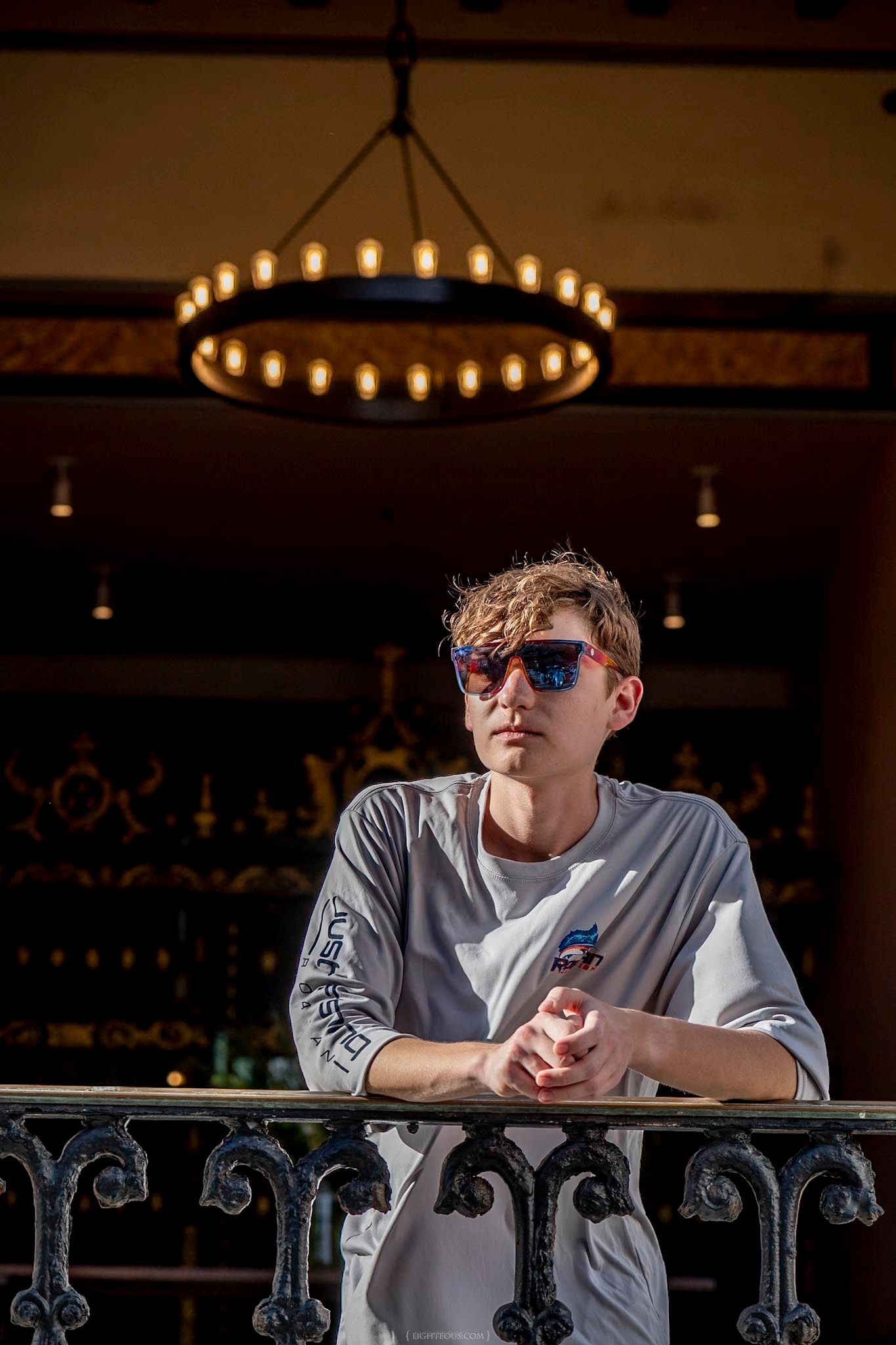 Young man in sunglasses and white t-shirt leaning on ornate iron balcony railing beneath a circular chandelier.