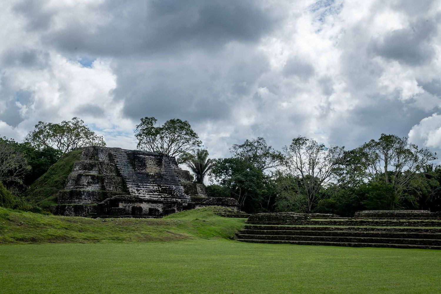 Ancient Mayan pyramids and structures rising from green fields under dramatic cloudy skies.