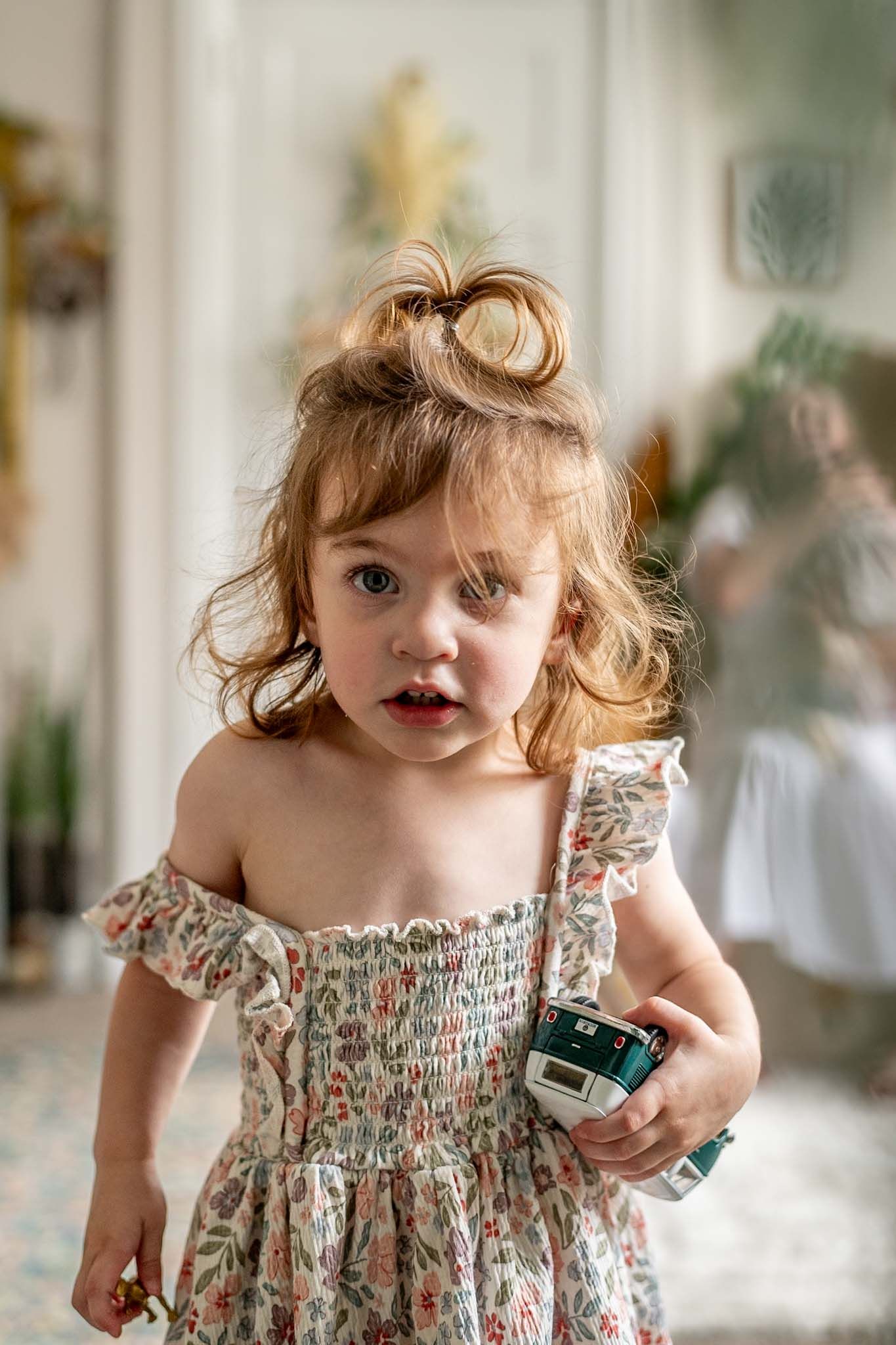 A toddler in a smocked floral dress plays expressively in a bright indoor setting.