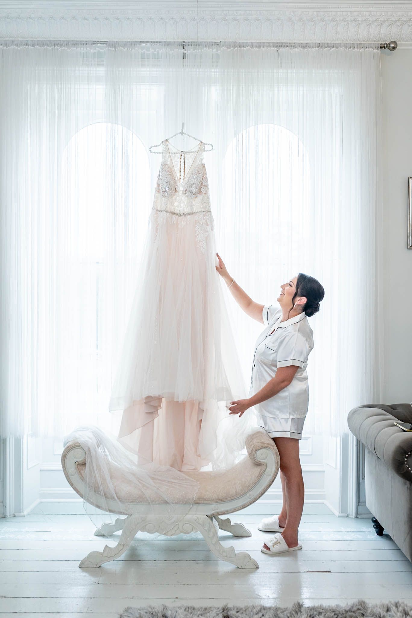 Bride admiring her elegant lace wedding dress hanging by a bright window in a white bridal suite.