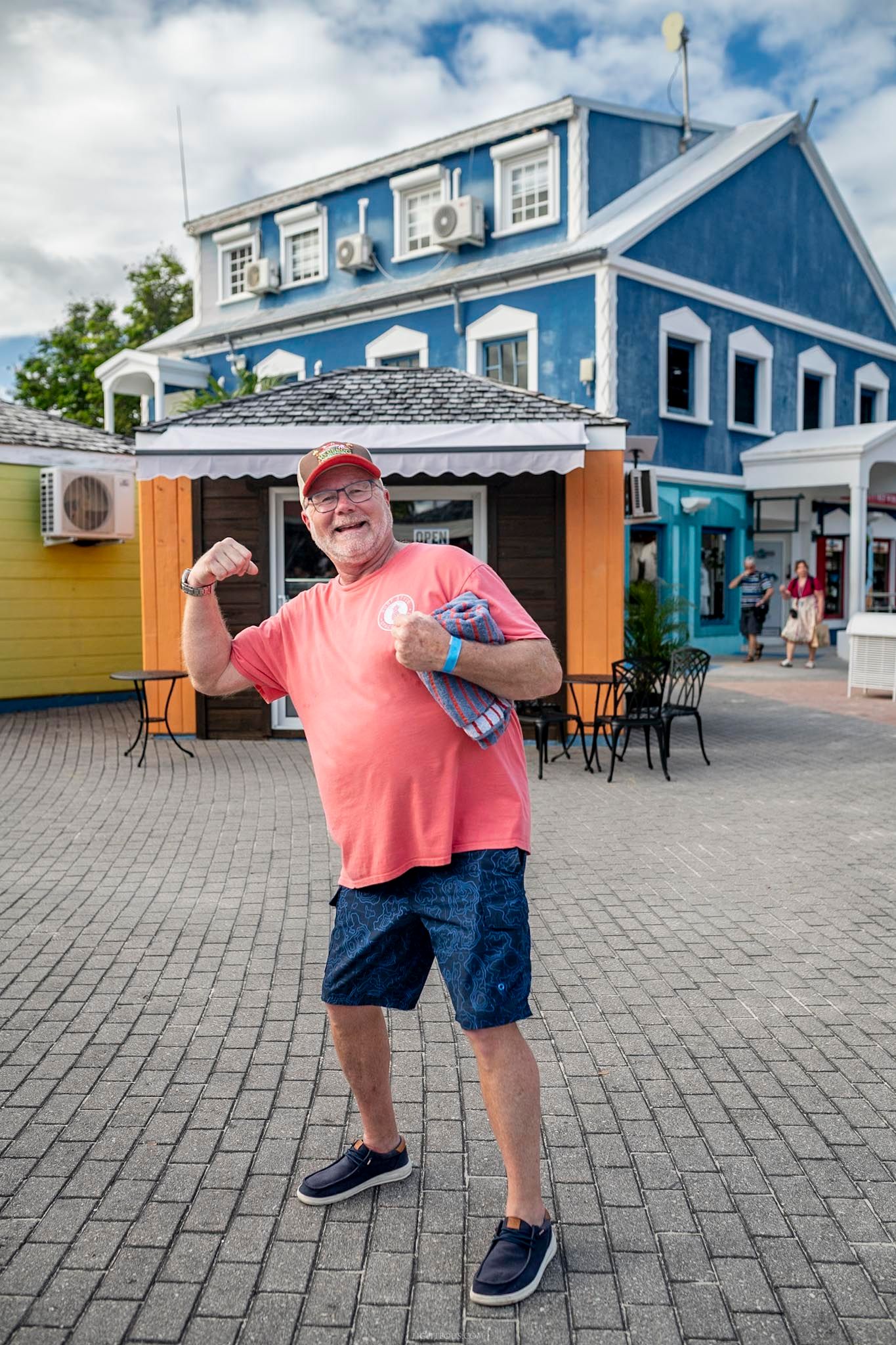 Happy senior man in pink shirt flexing his arm in front of a blue building at a tropical shopping area.