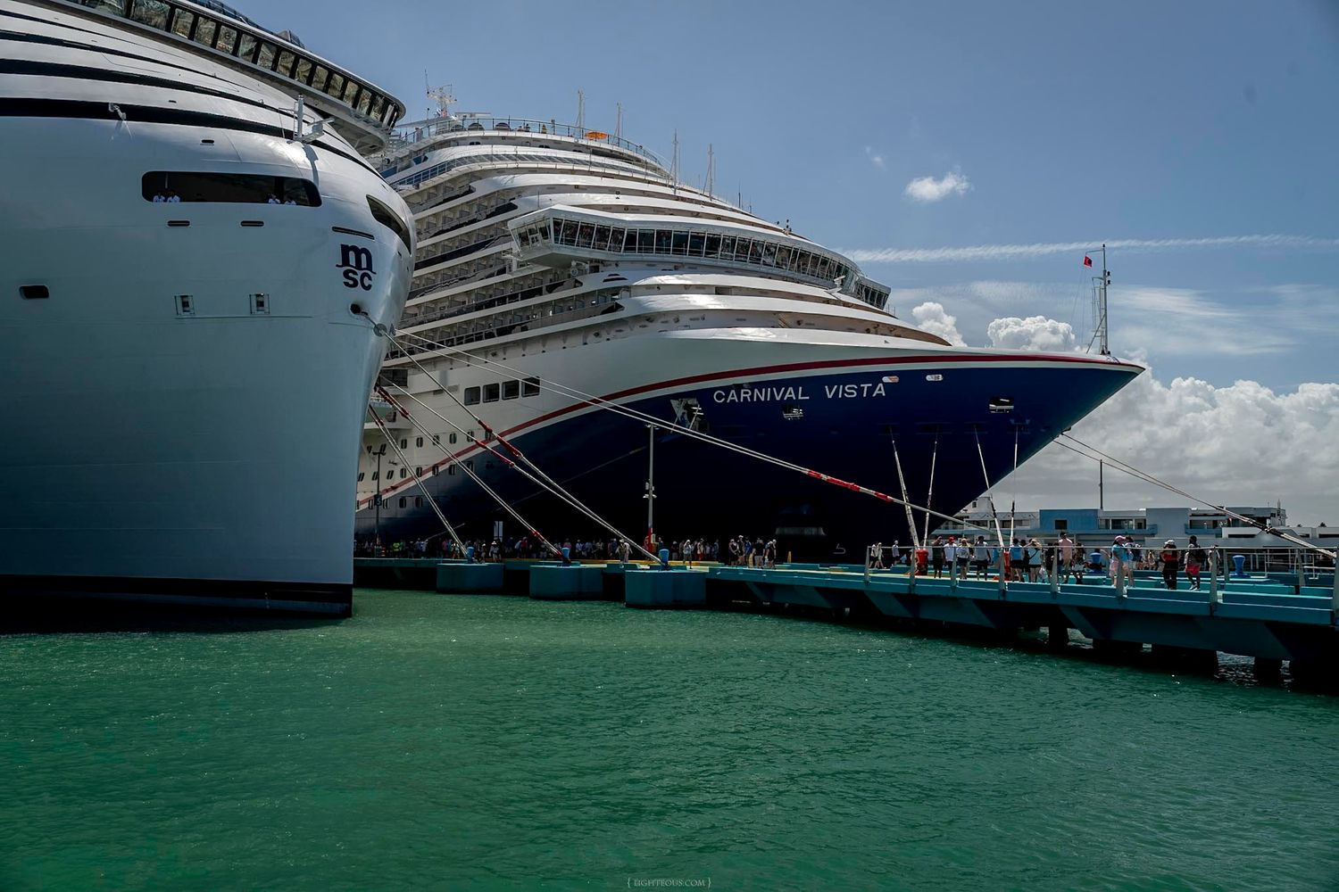 Carnival Vista cruise ship docked at a tropical port alongside another large cruise liner under partly cloudy skies.