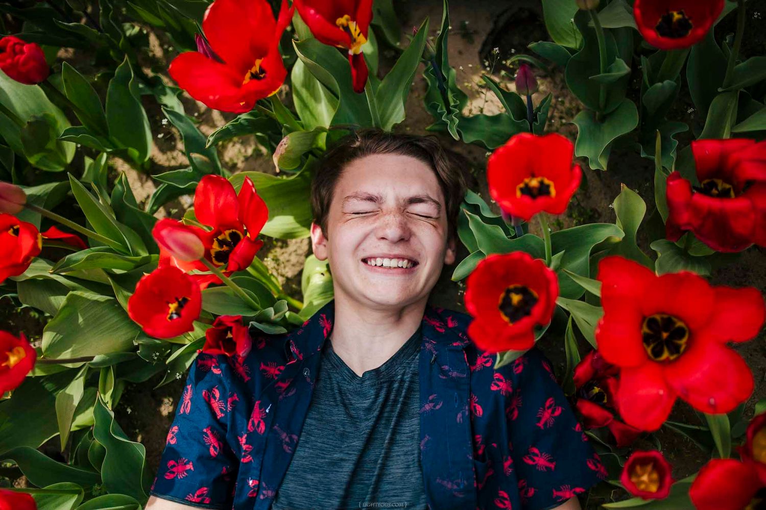 A joyful young person lying among vibrant red tulips, smiling with eyes closed in a lush garden.