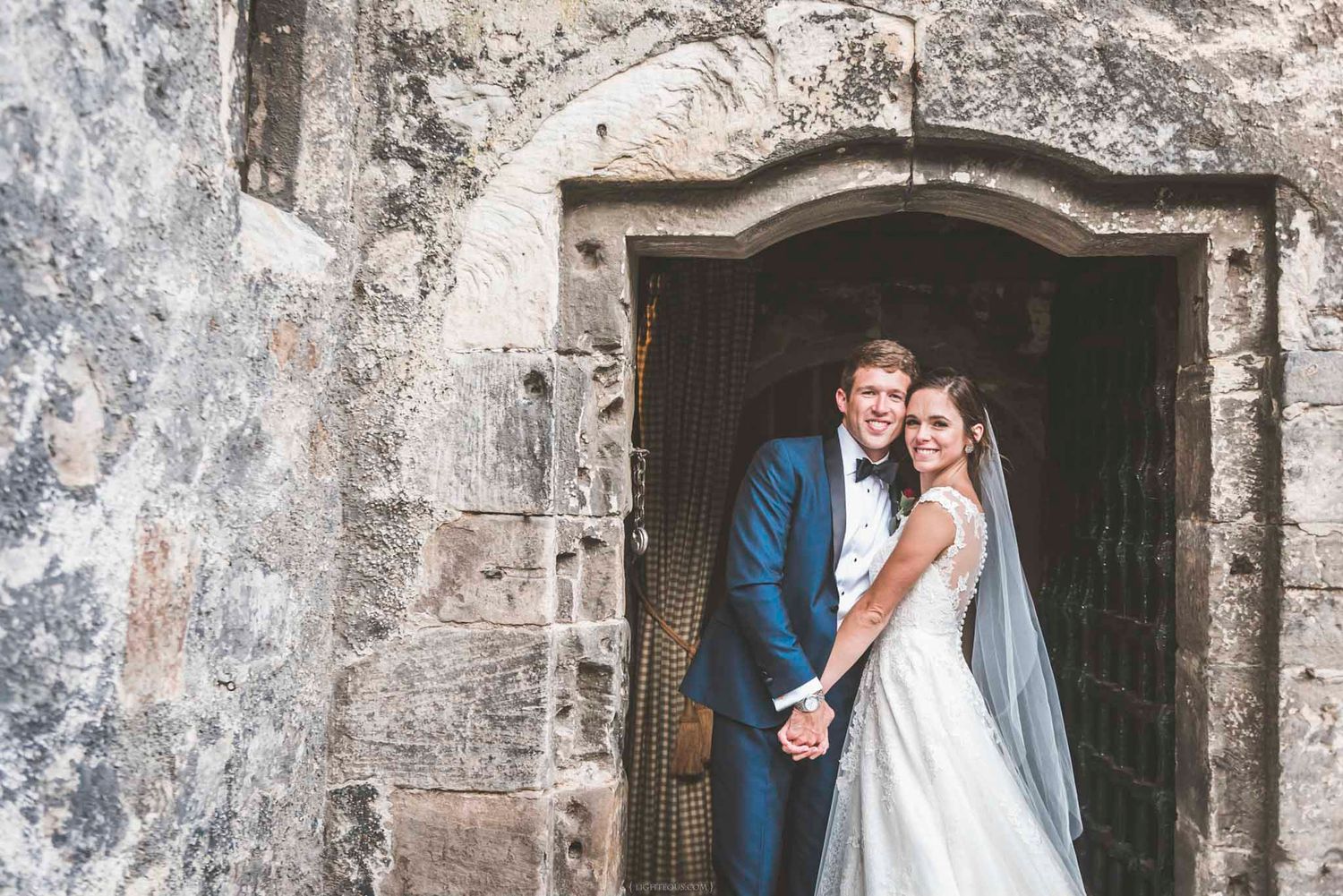 Bride and groom posing happily in a historic stone doorway, groom in navy suit, bride in white gown.
