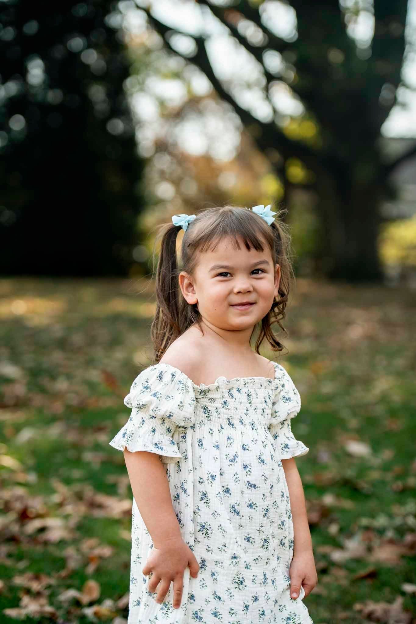 A smiling young girl with pigtails wearing a white floral off-shoulder dress stands outdoors in a park.