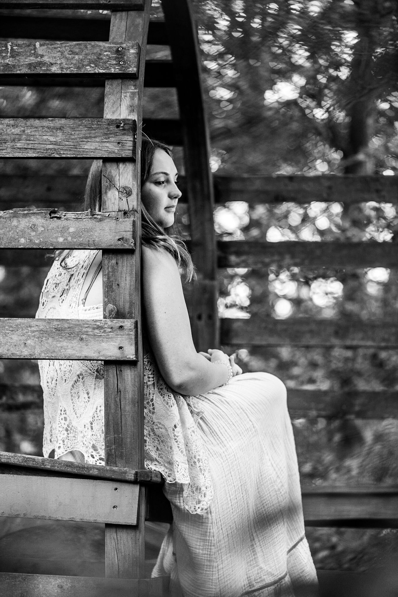 Black and white photo of a young woman in a white dress sitting on rustic wooden stairs surrounded by trees.