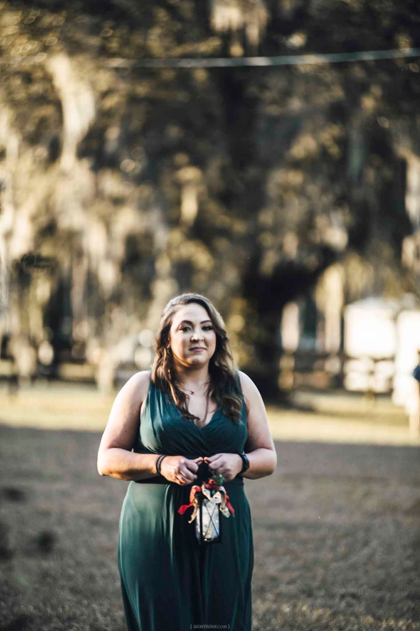 Bridesmaid in elegant green gown holds a floral bouquet outdoors among moss-draped trees.