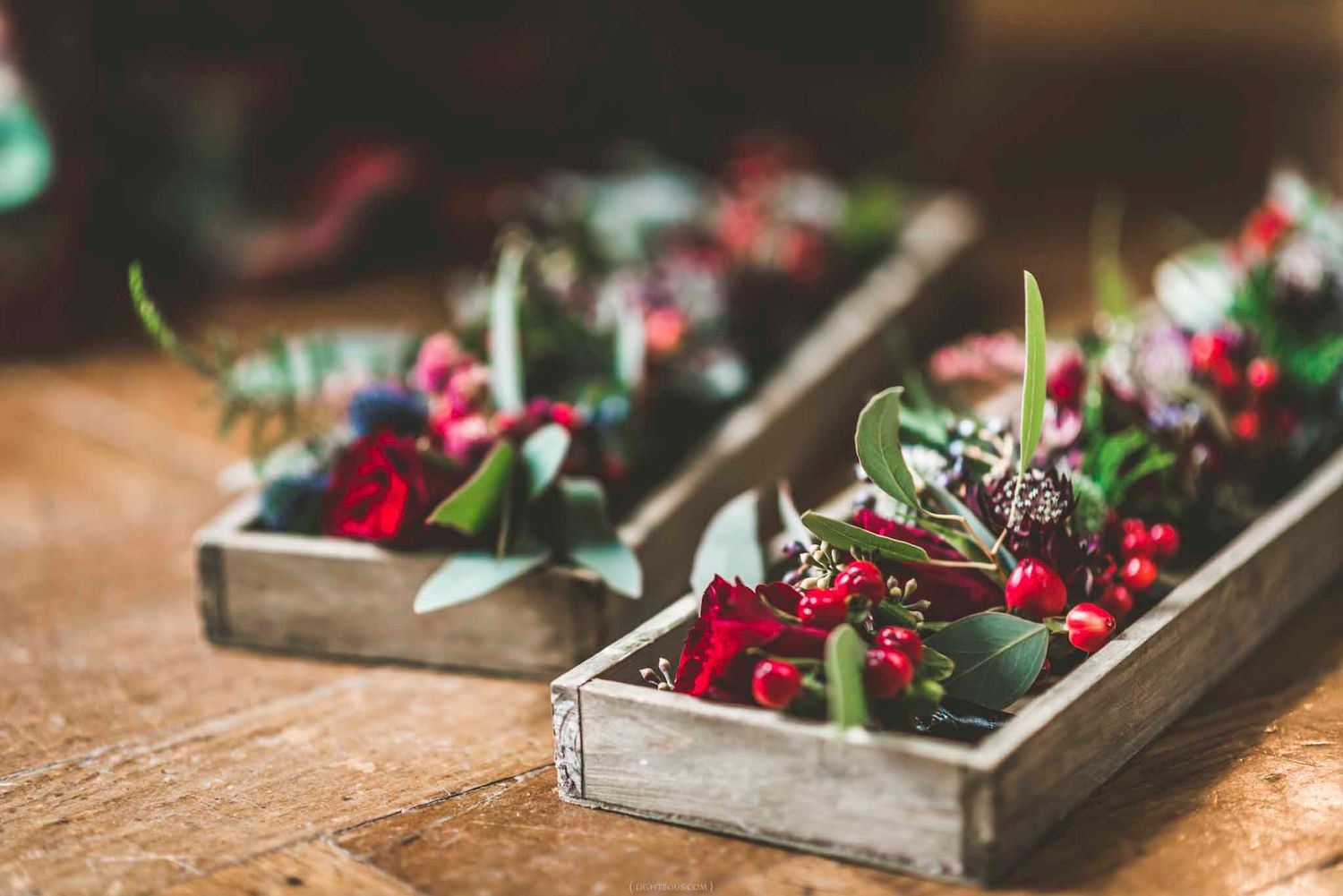 Rustic wooden boxes filled with red berries, dark flowers, and green foliage centerpieces on a wooden table.