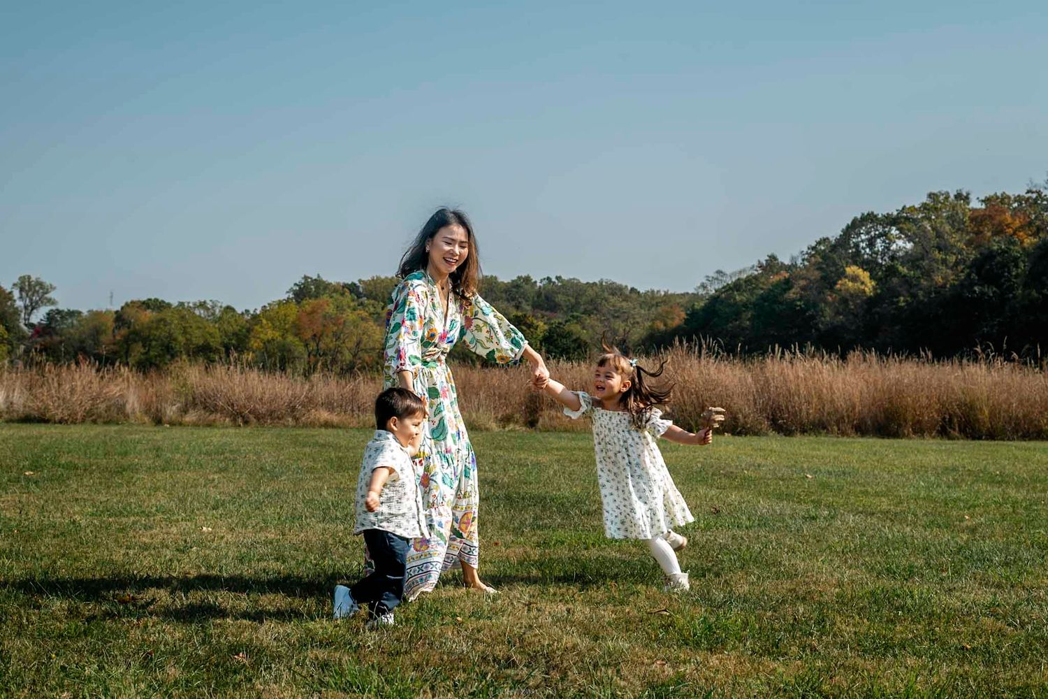 A mother in a floral dress plays with her two young children on a green field under a clear blue sky.