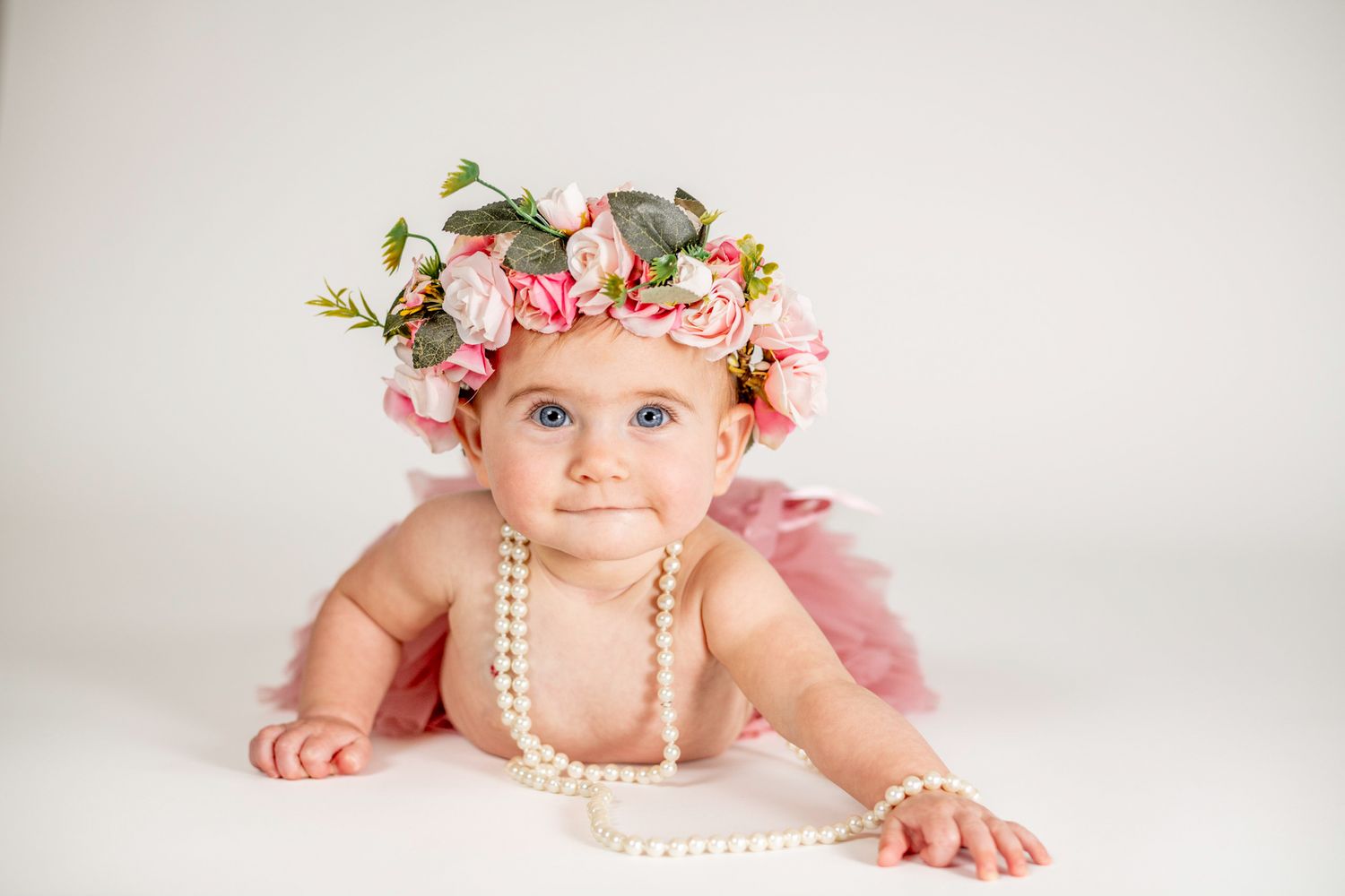 Adorable baby girl wearing a pink floral crown and pearl necklace, posing on a white background in a pink tutu.