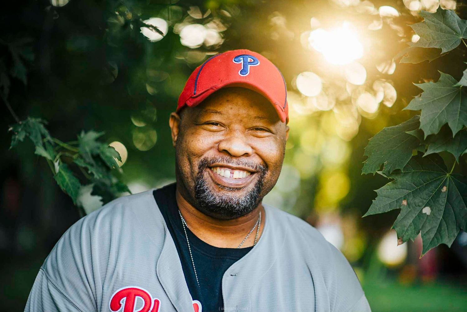 Smiling man wearing a red Philadelphia Phillies cap and jersey outdoors with green foliage background.