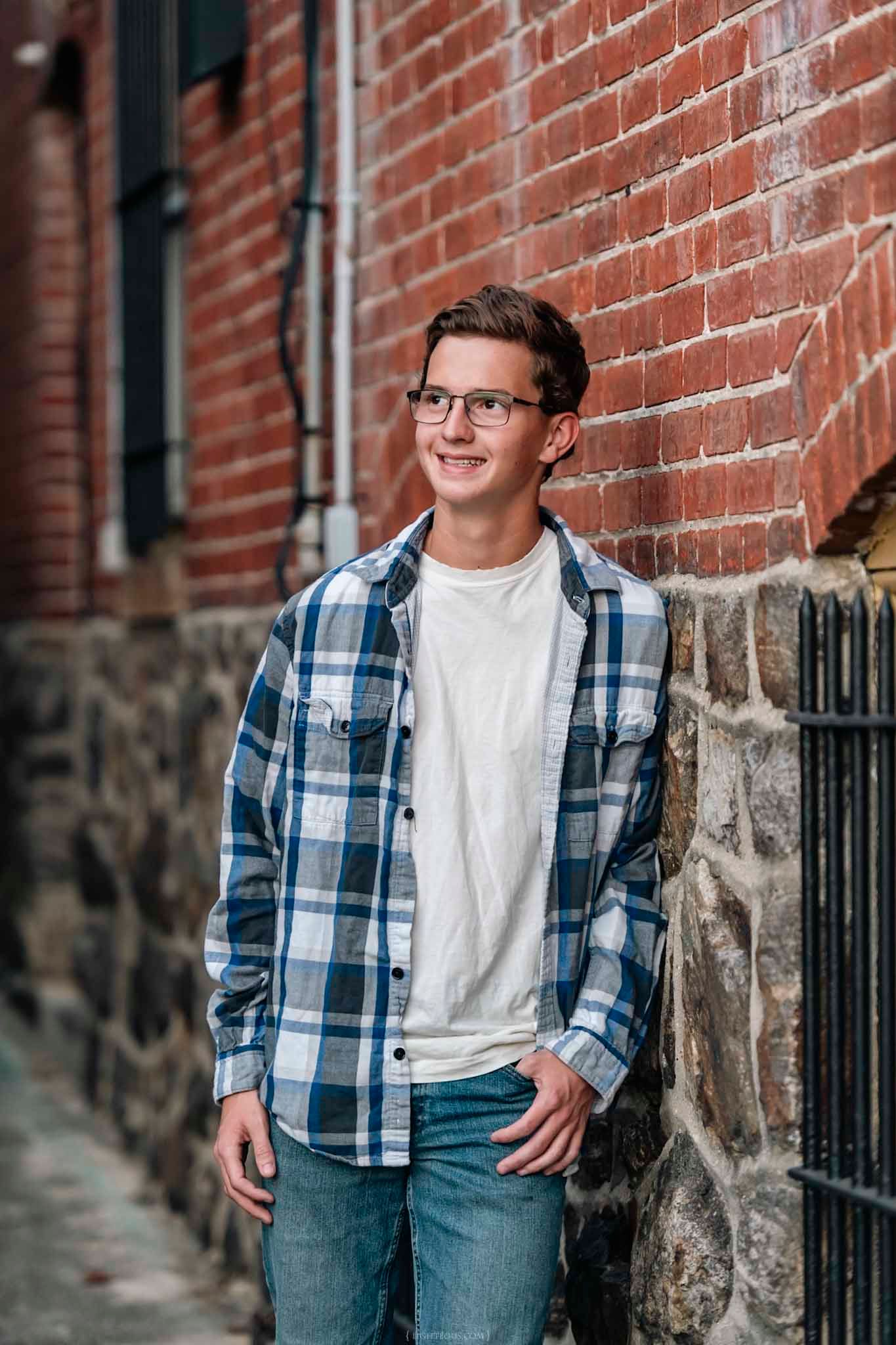 Young man in plaid shirt and glasses smiling while leaning against a brick wall outdoors.