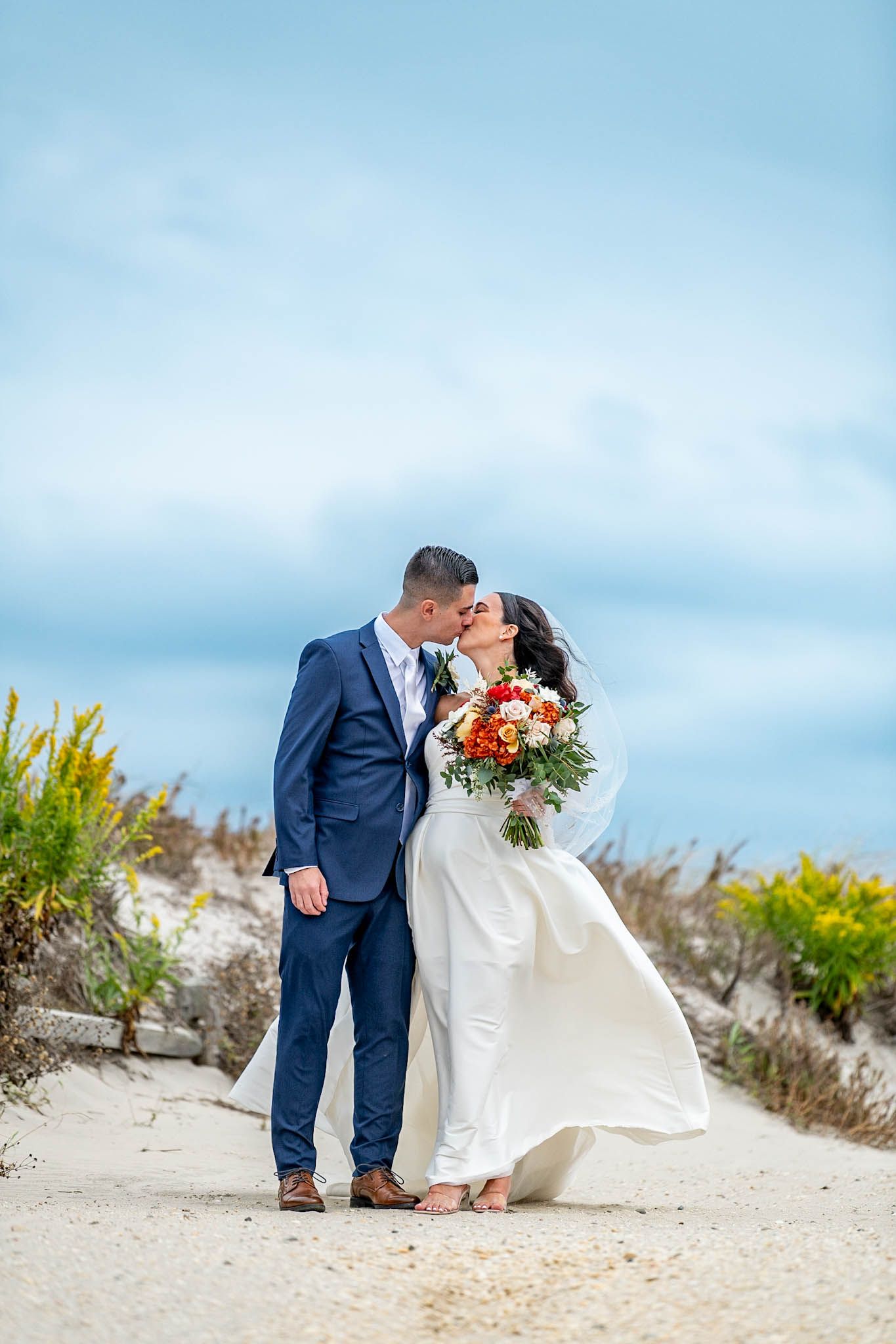 Bride and groom kissing on sandy beach path, bride holding colorful bouquet, groom in navy suit, blue sky backdrop.