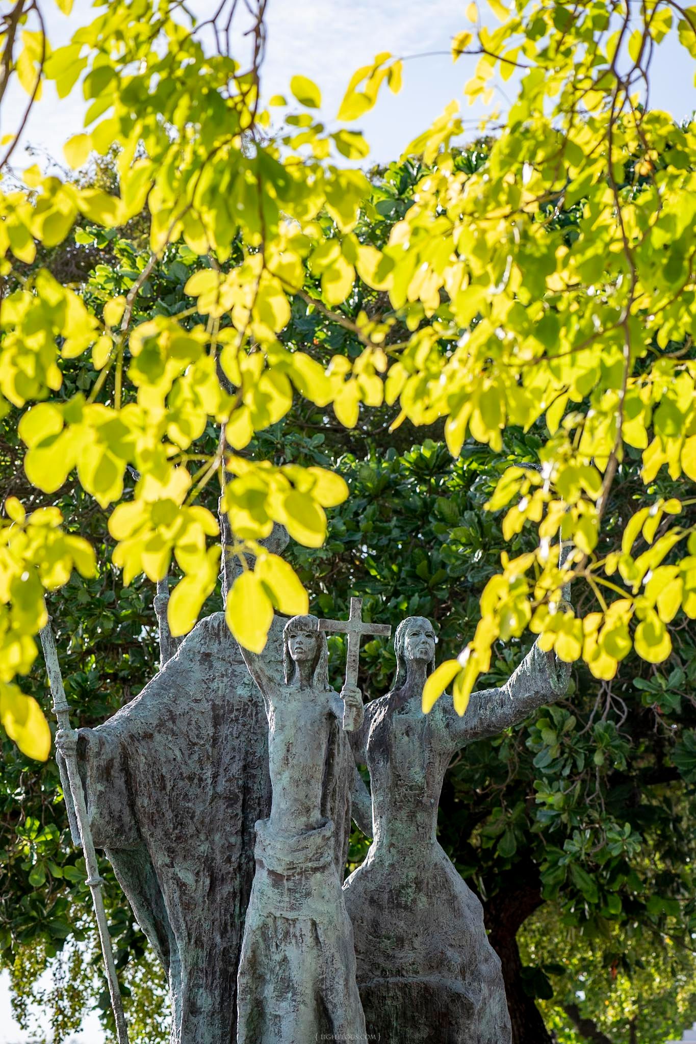 Bronze statue of two figures holding a cross, framed by vibrant yellow-green leaves and lush trees in sunlight.