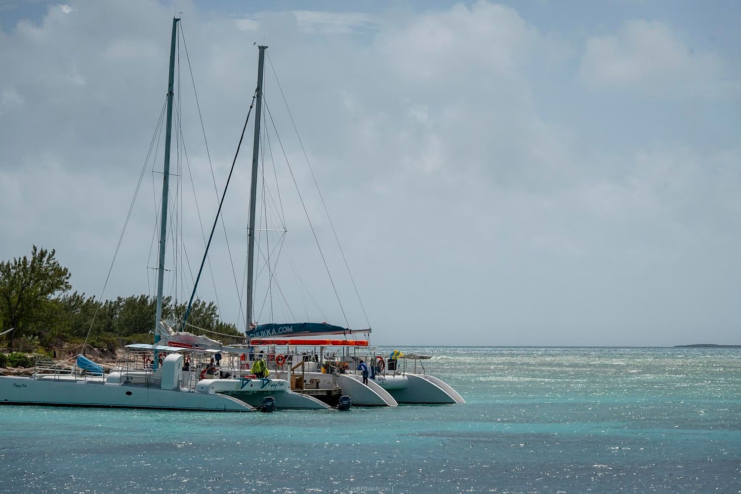 Catamaran sailboats docked near a tropical island shore with turquoise calm waters under a cloudy sky.