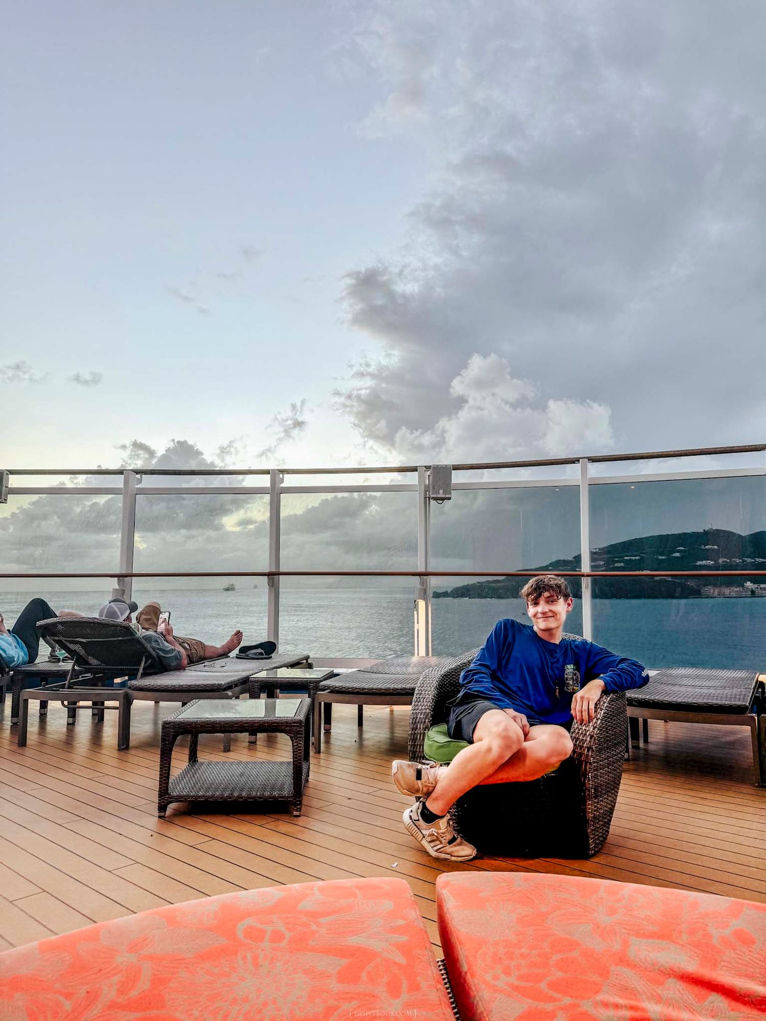 Young man in blue hoodie relaxing on cruise ship deck with panoramic ocean and island views at dusk.