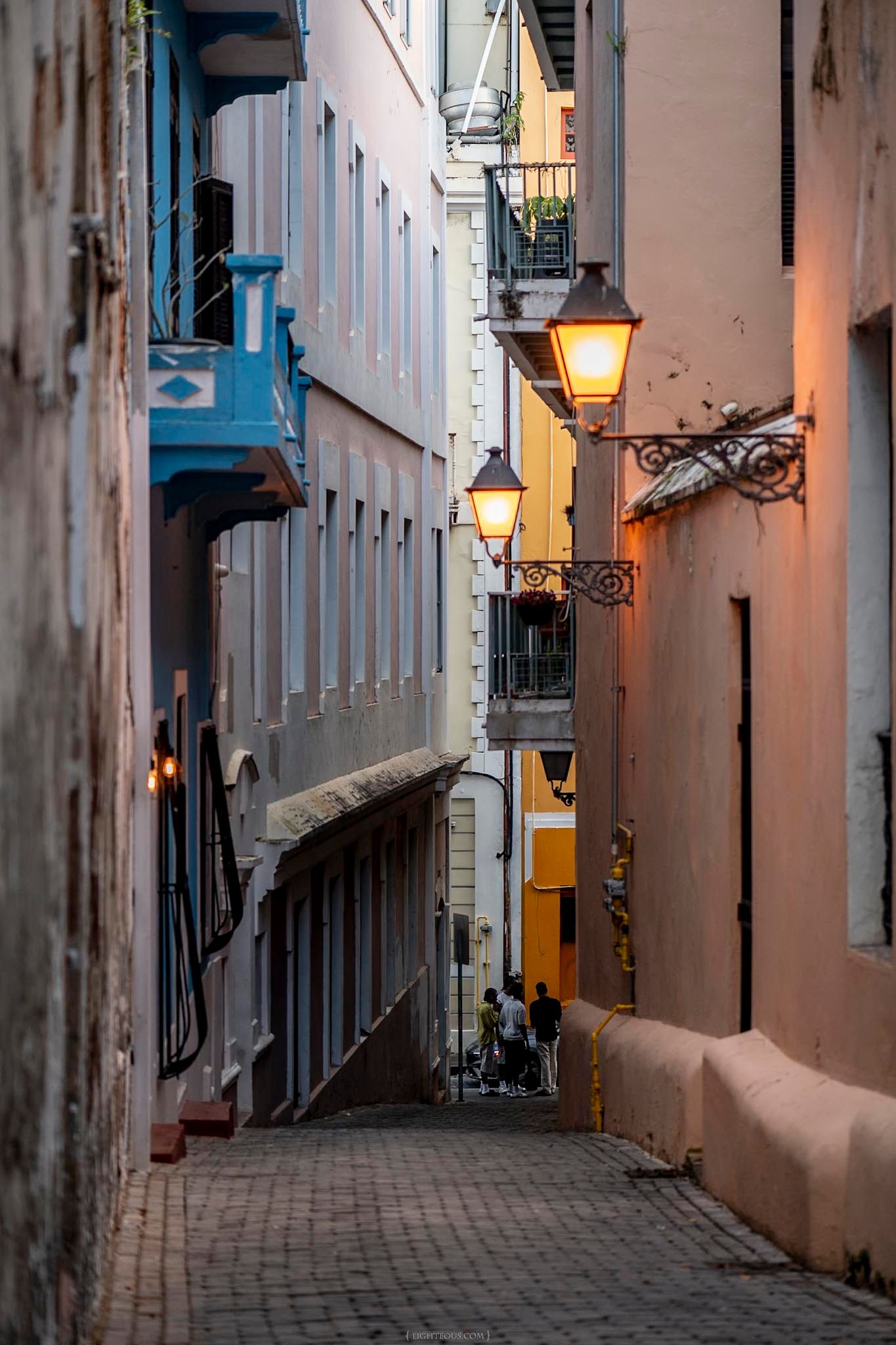 Narrow cobblestone alley in Old San Juan illuminated by warm vintage street lamps at dusk.