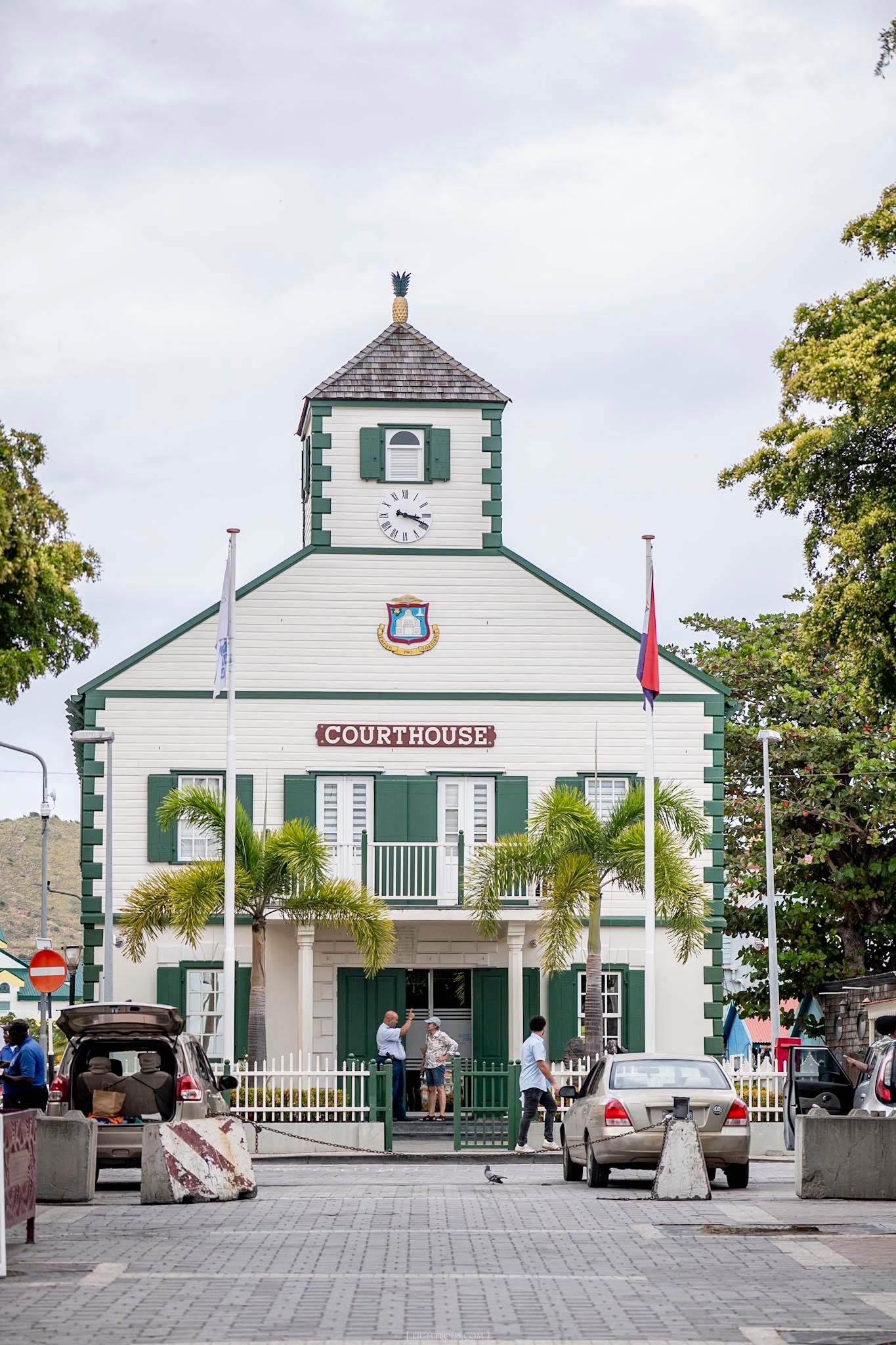 White colonial courthouse building with green shutters, clock tower, palm trees, and flags in Philipsburg, Sint Maarten.