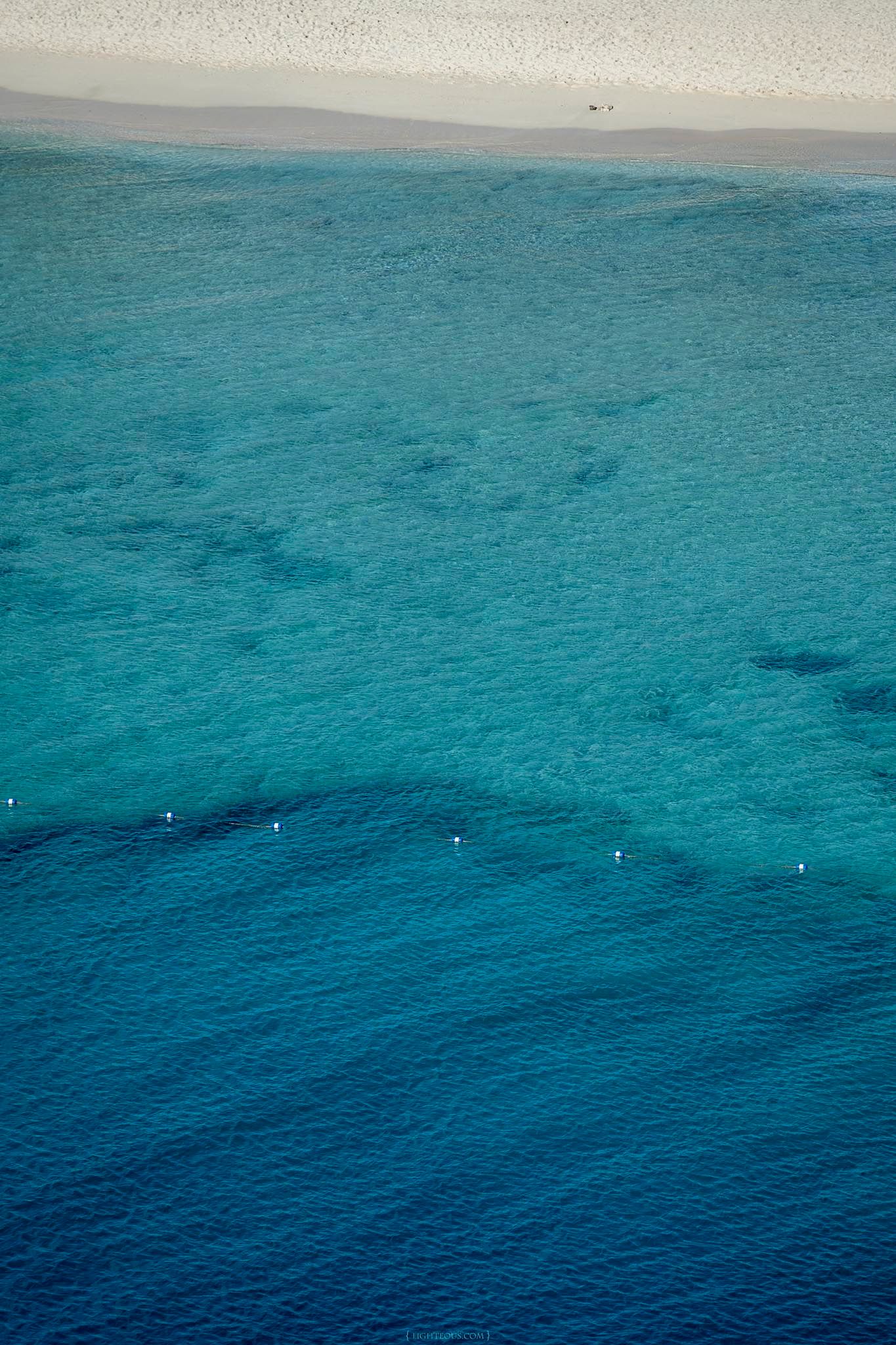 Aerial view of crystal clear turquoise ocean water with a white sandy beach shoreline in the background.