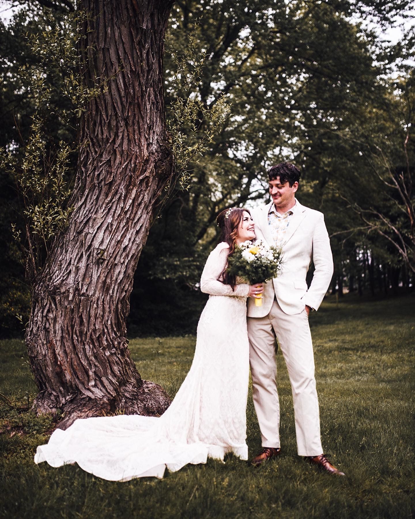 A bride in a white lace gown and groom in a tan suit stand by a large tree, sharing a romantic moment outdoors.