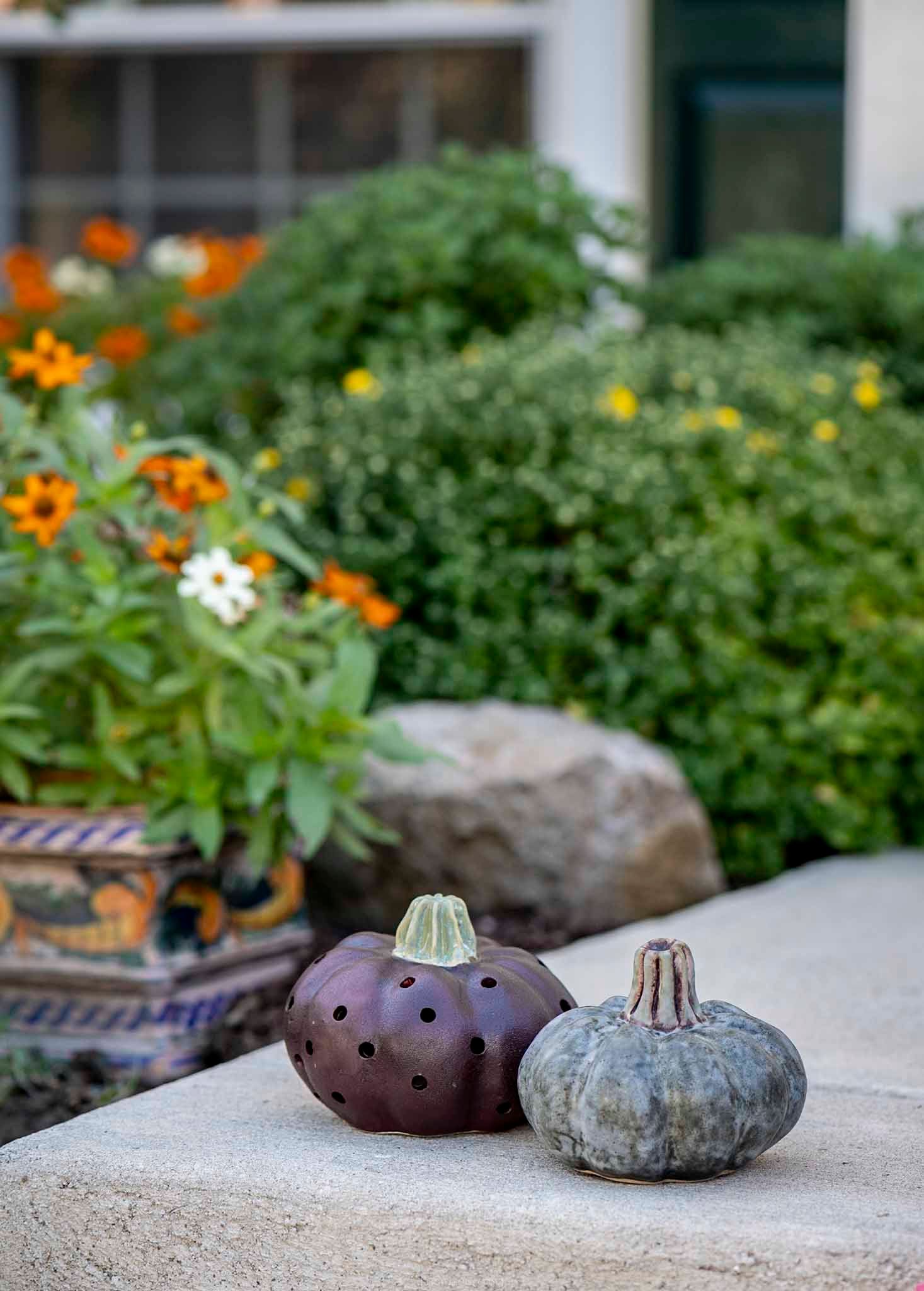 Two ceramic pumpkin figurines on a stone ledge with colorful garden flowers and greenery in the background.