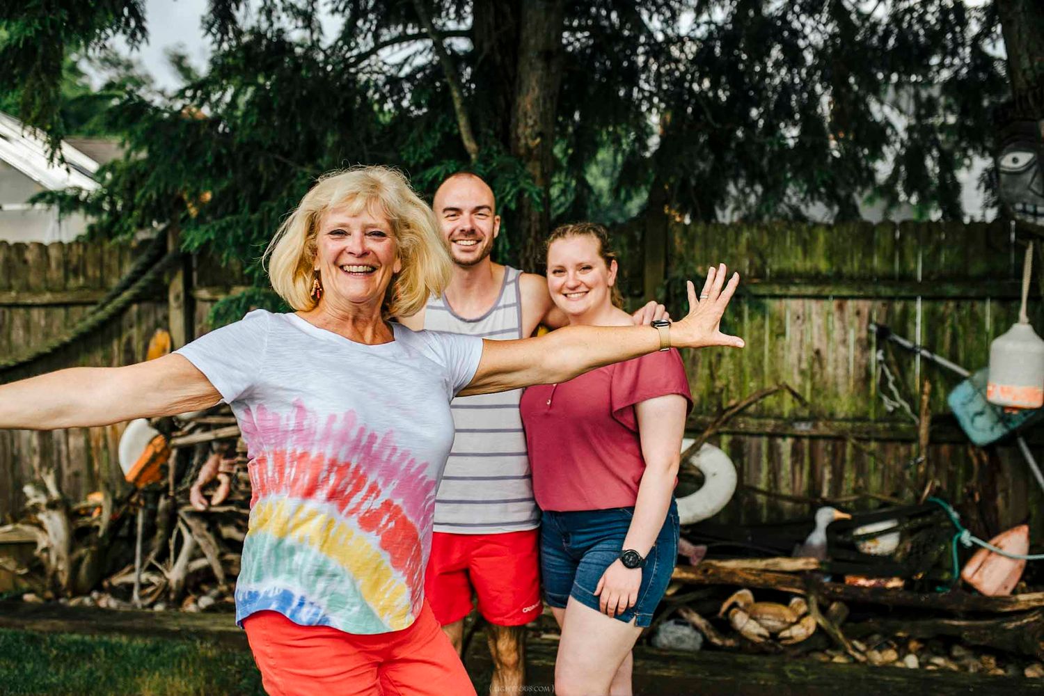 Three people smiling and posing with arms outstretched in a backyard, wearing casual summer clothing.