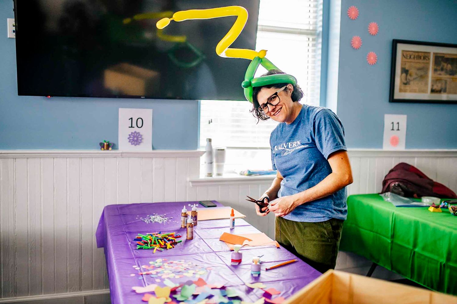 A woman wearing a green hat crafts at a colorful table during a festive indoor activity event.