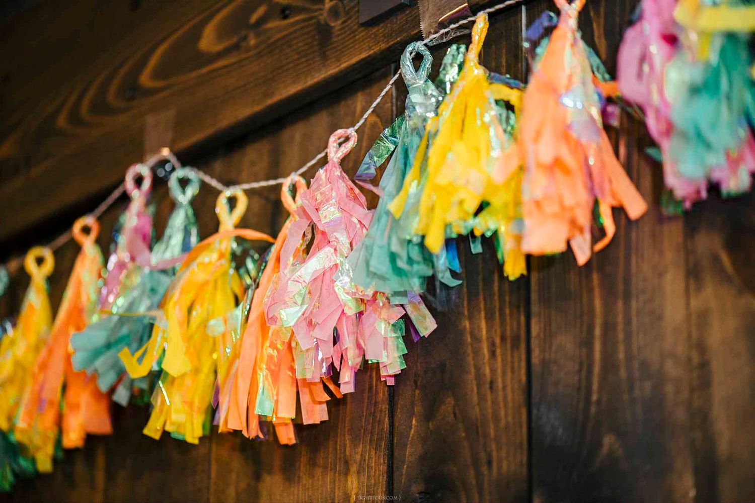 Colorful tissue paper tassel garland in pink, yellow, orange, and teal hanging against a rustic wood wall.