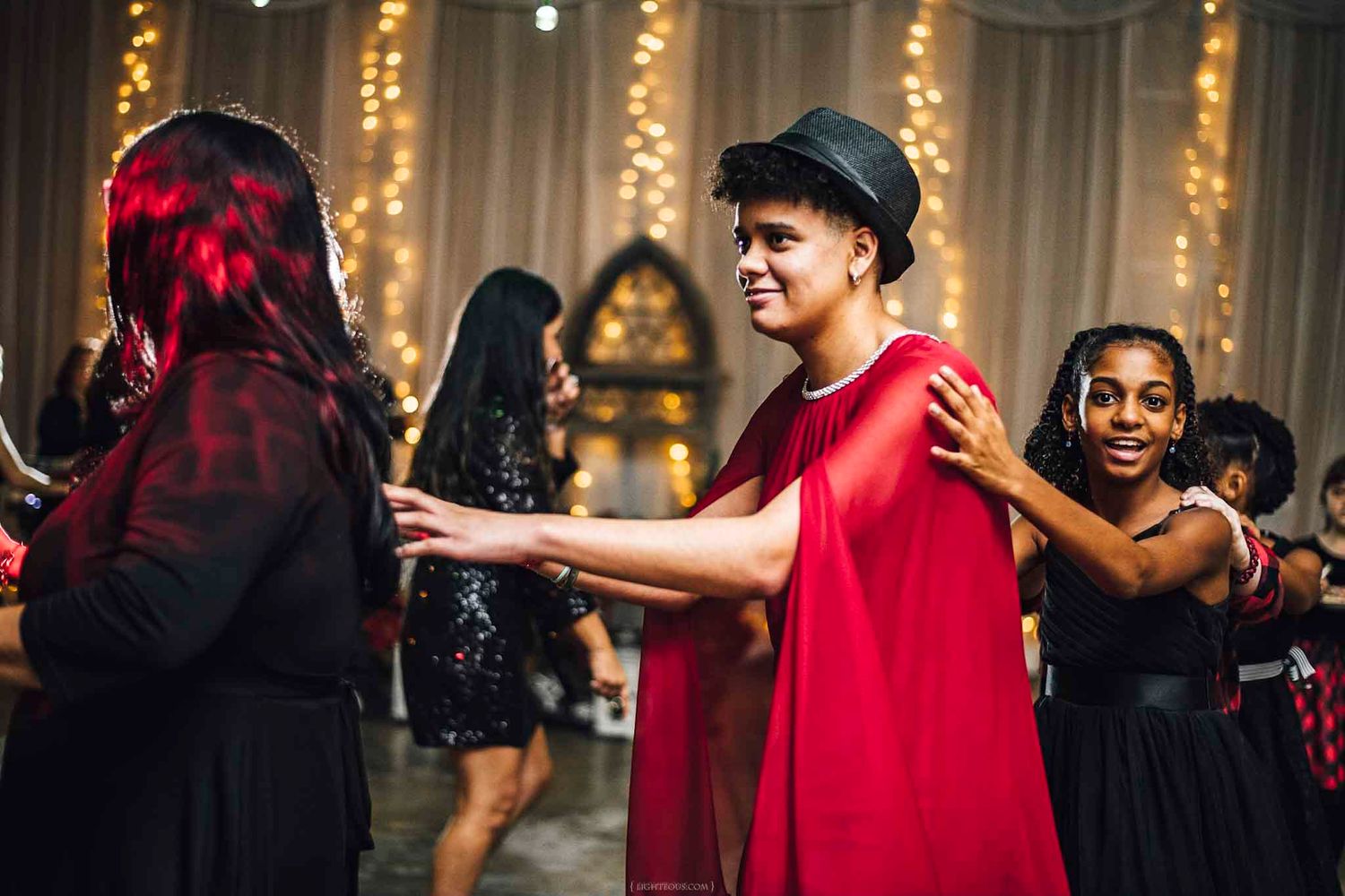 Teen in red cape and black hat dances with friends at a festive school dance event with string lights.