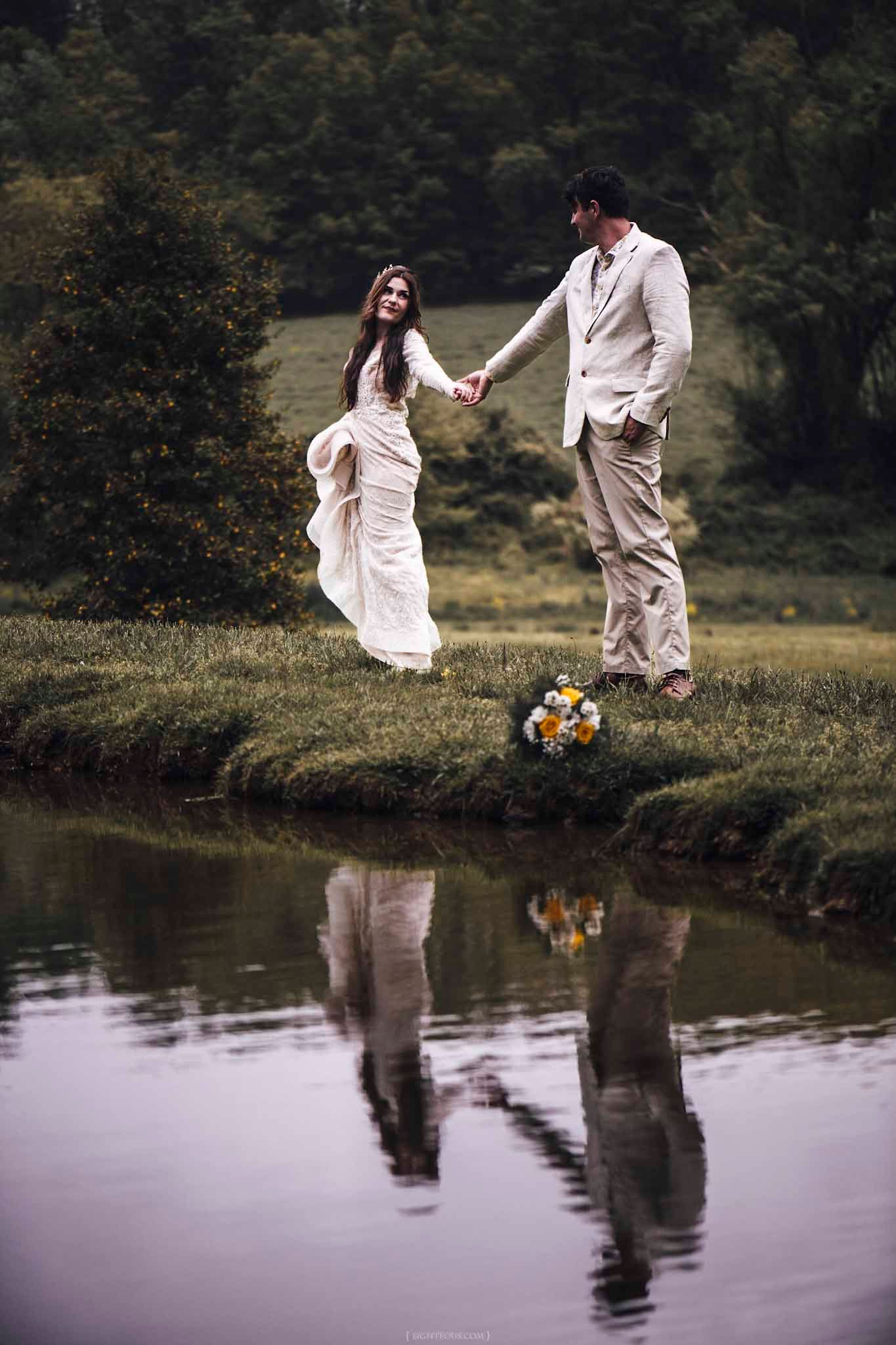 Bride and groom holding hands by a reflective pond, with a floral bouquet on the grass and lush green trees behind.